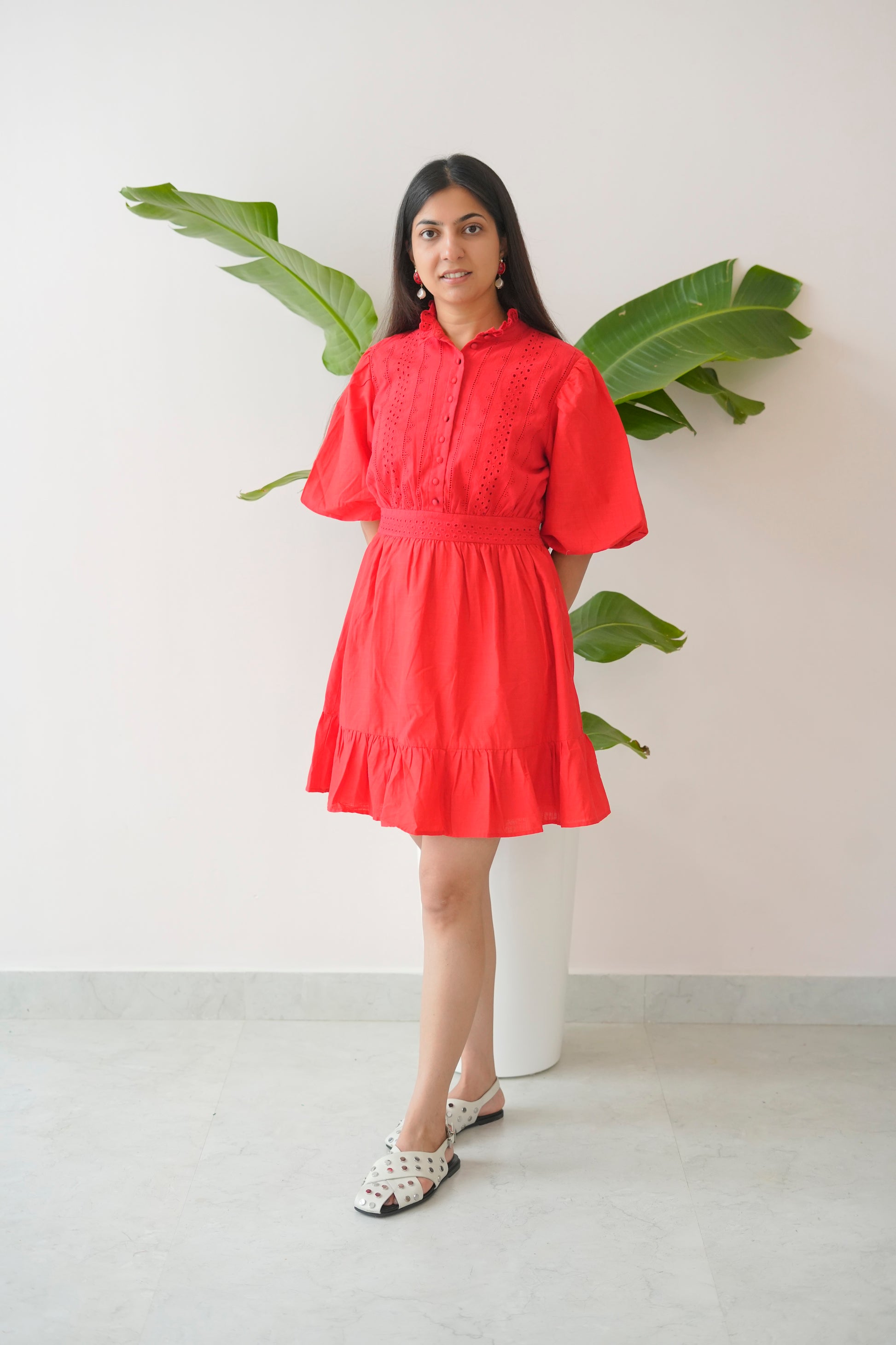 Woman wearing a red dress standing in front of a white wall with green leaves.