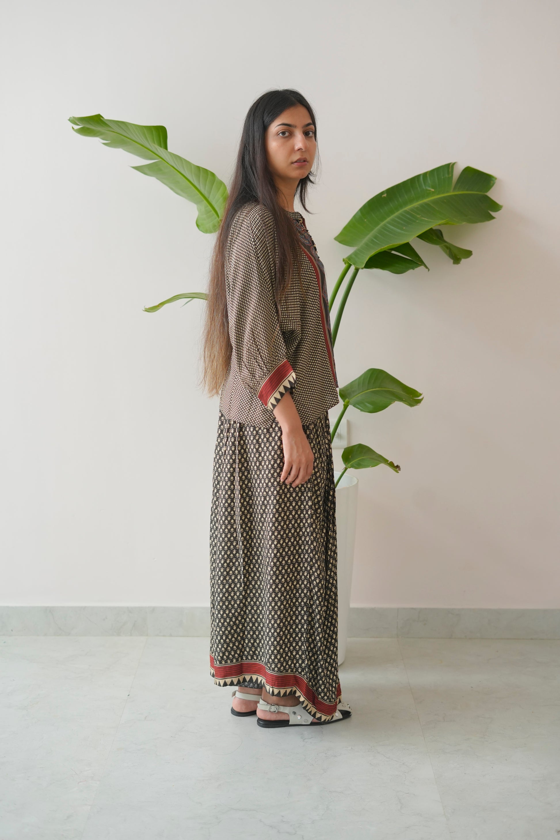 Woman in a patterned dress standing next to a large plant against a plain wall.