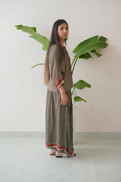 Woman in a patterned dress standing next to a large plant against a plain wall.