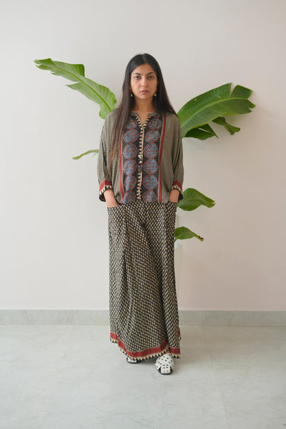 Woman wearing a patterned outfit standing in front of a white wall with green leaves.