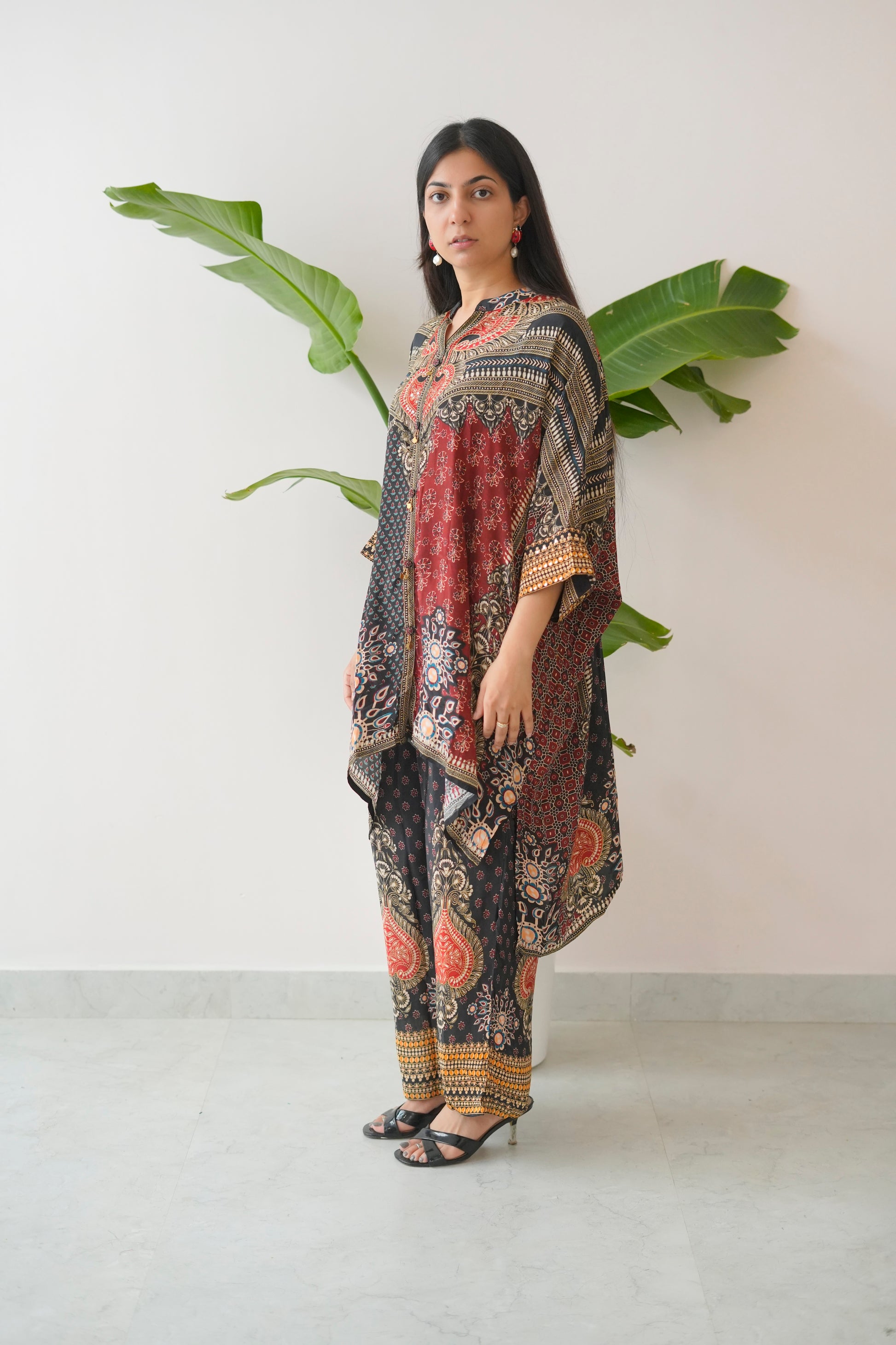 Woman wearing a patterned outfit standing against a white wall with a plant.