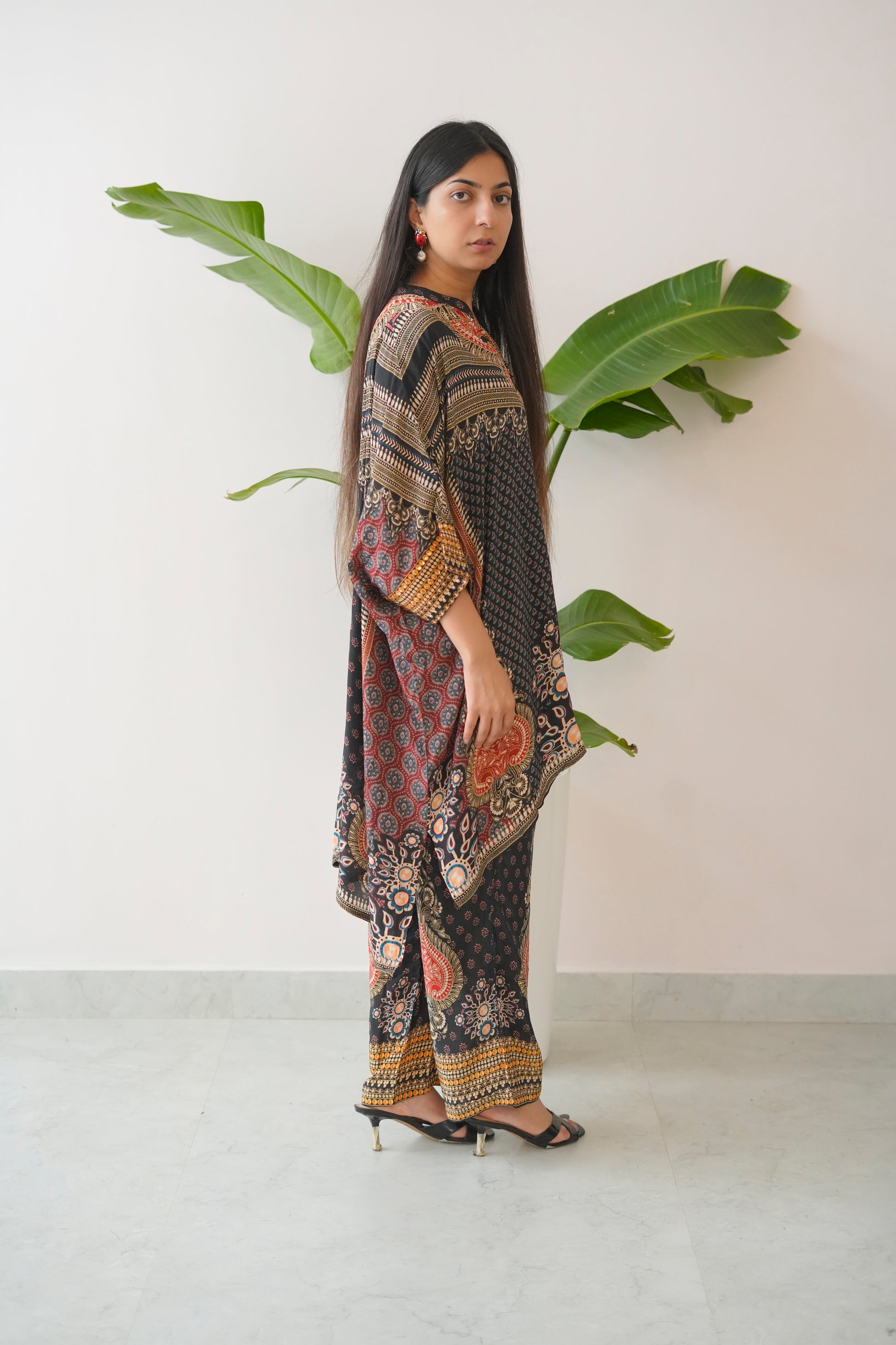 Woman wearing a patterned outfit standing next to a plant against a white wall.