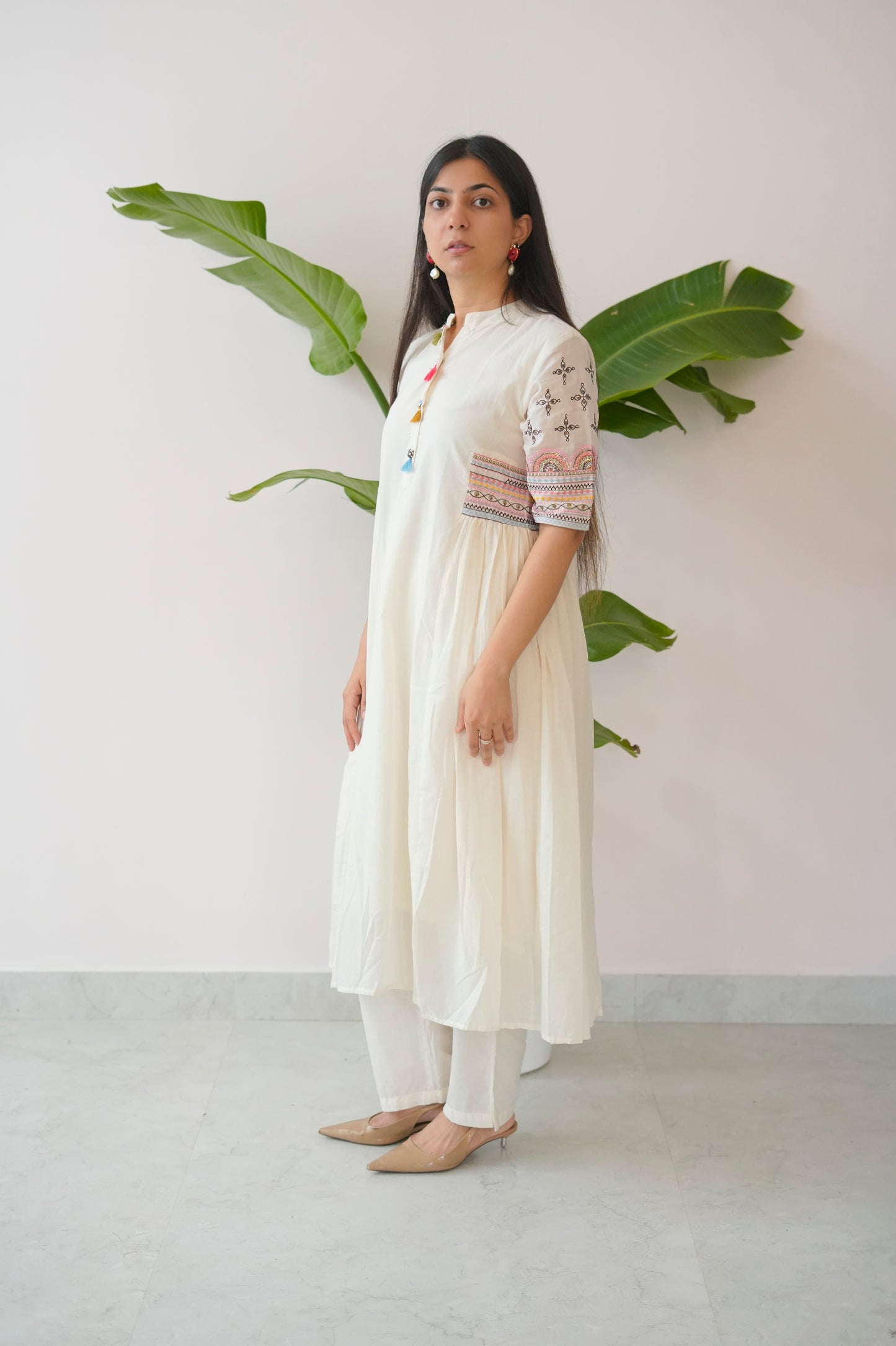 Woman wearing a white outfit with embroidered details standing against a plain background with a plant.