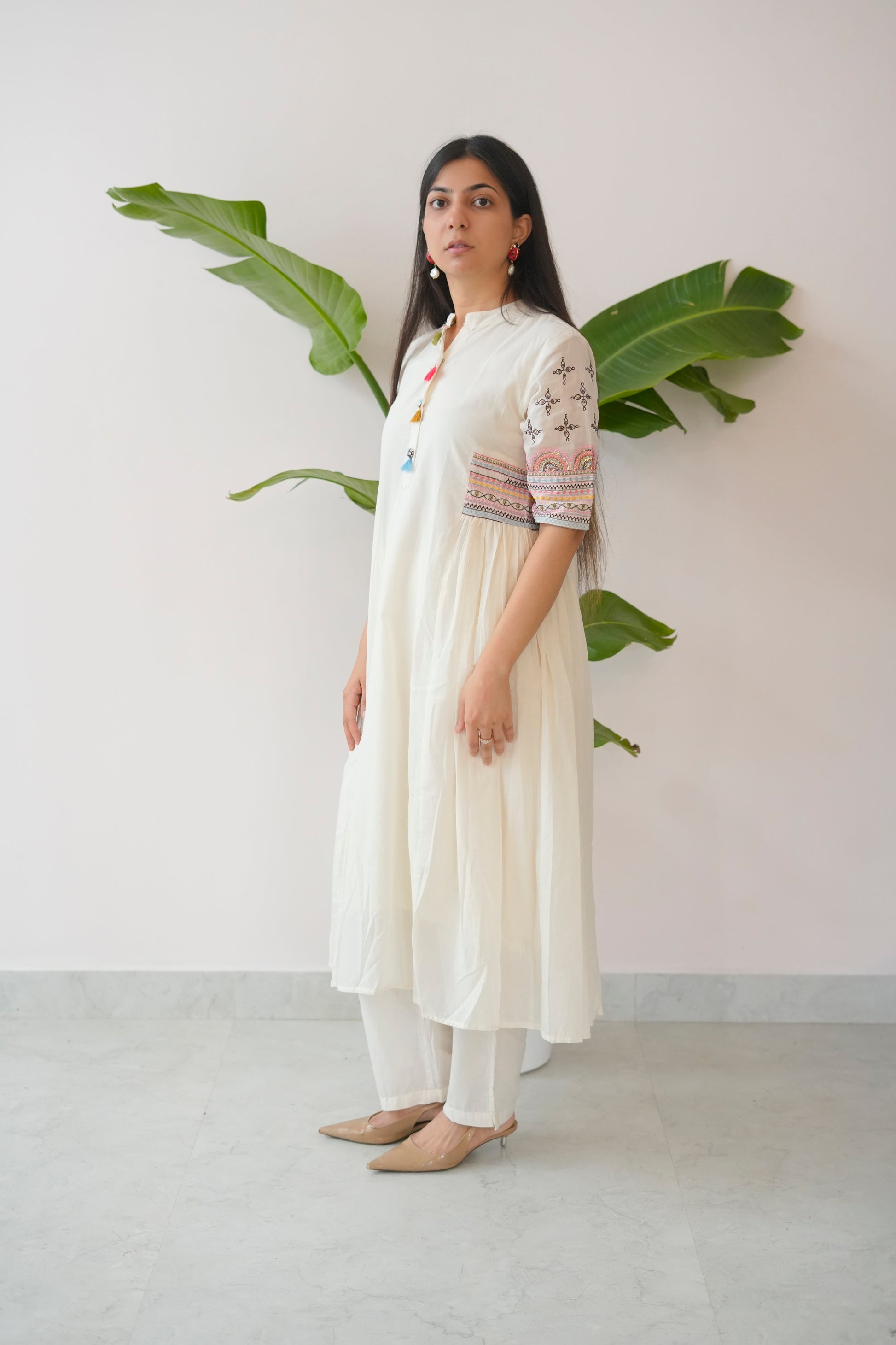 Woman wearing a white outfit with embroidered details standing against a plain background with a plant.