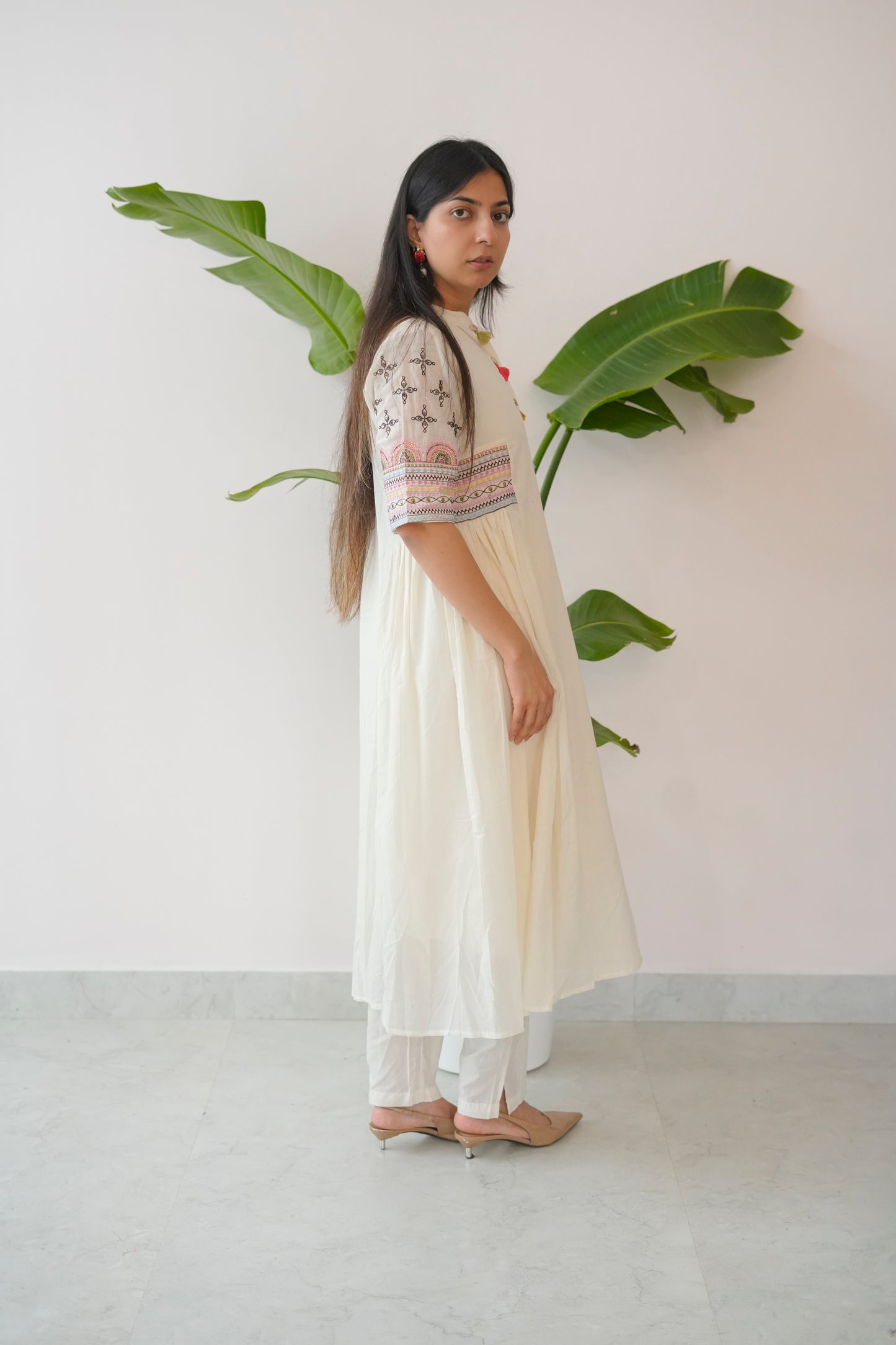 Woman in a white dress with floral patterns standing next to a plant against a plain wall.