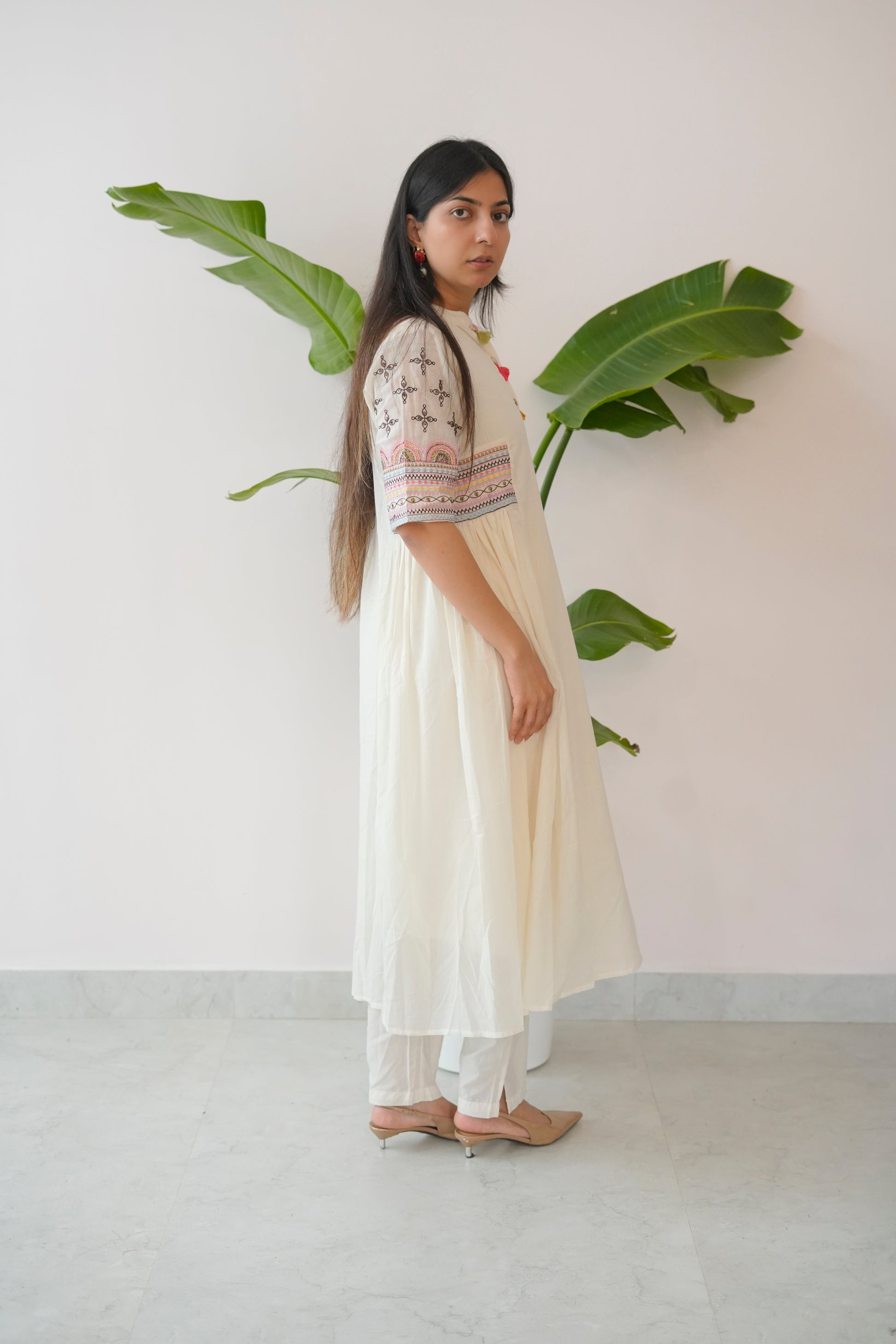 Woman in a white dress with floral patterns standing next to a plant against a plain wall.