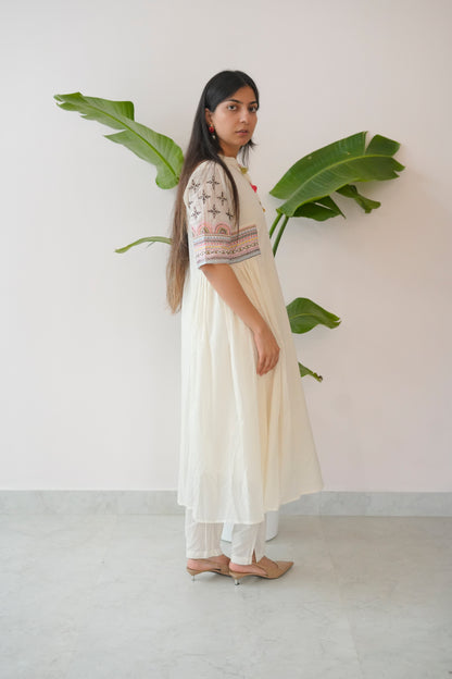 Woman in a white dress with floral patterns standing next to a plant against a plain wall.