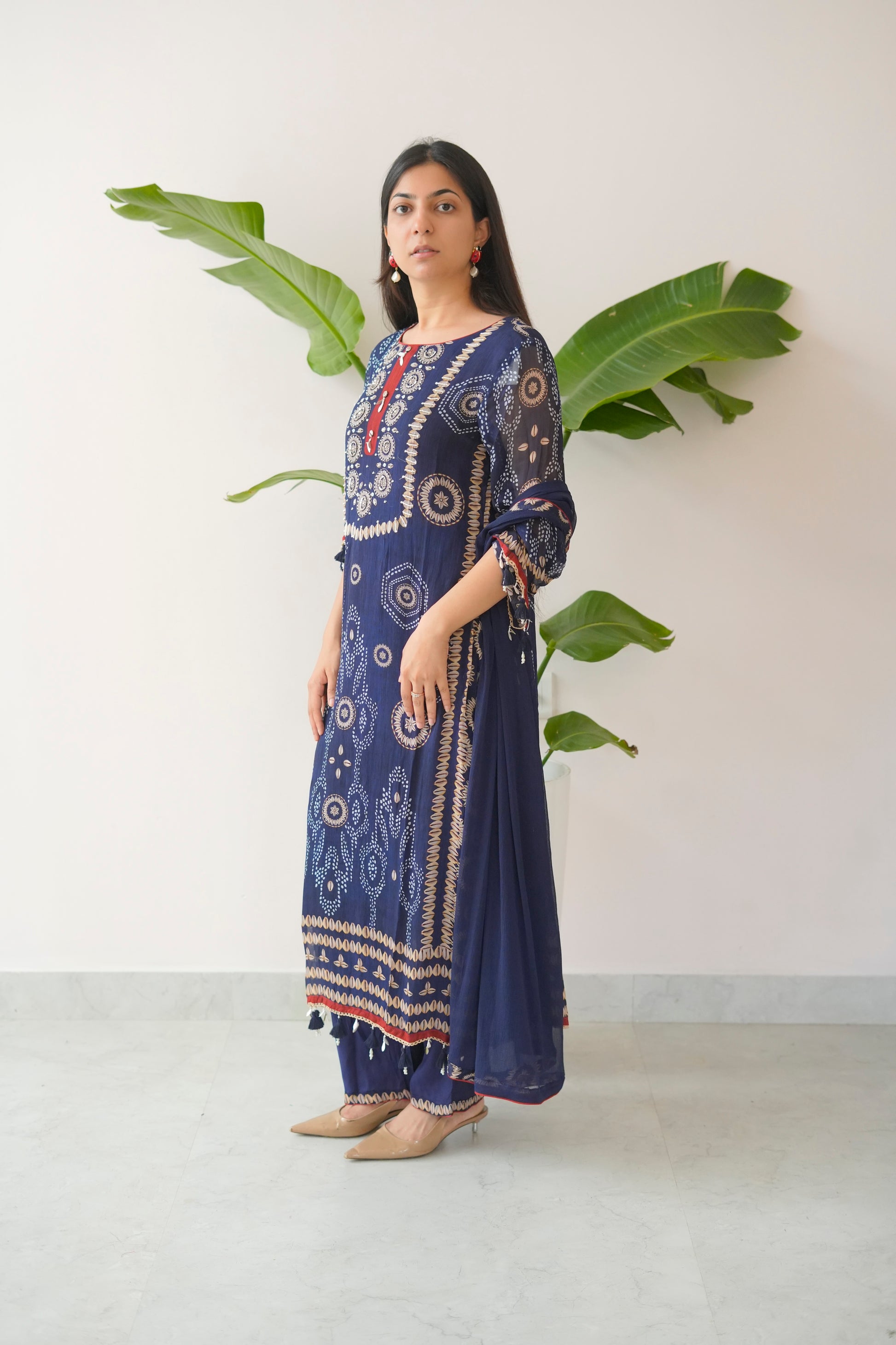 Woman wearing a blue patterned dress standing against a white wall with green leaves.