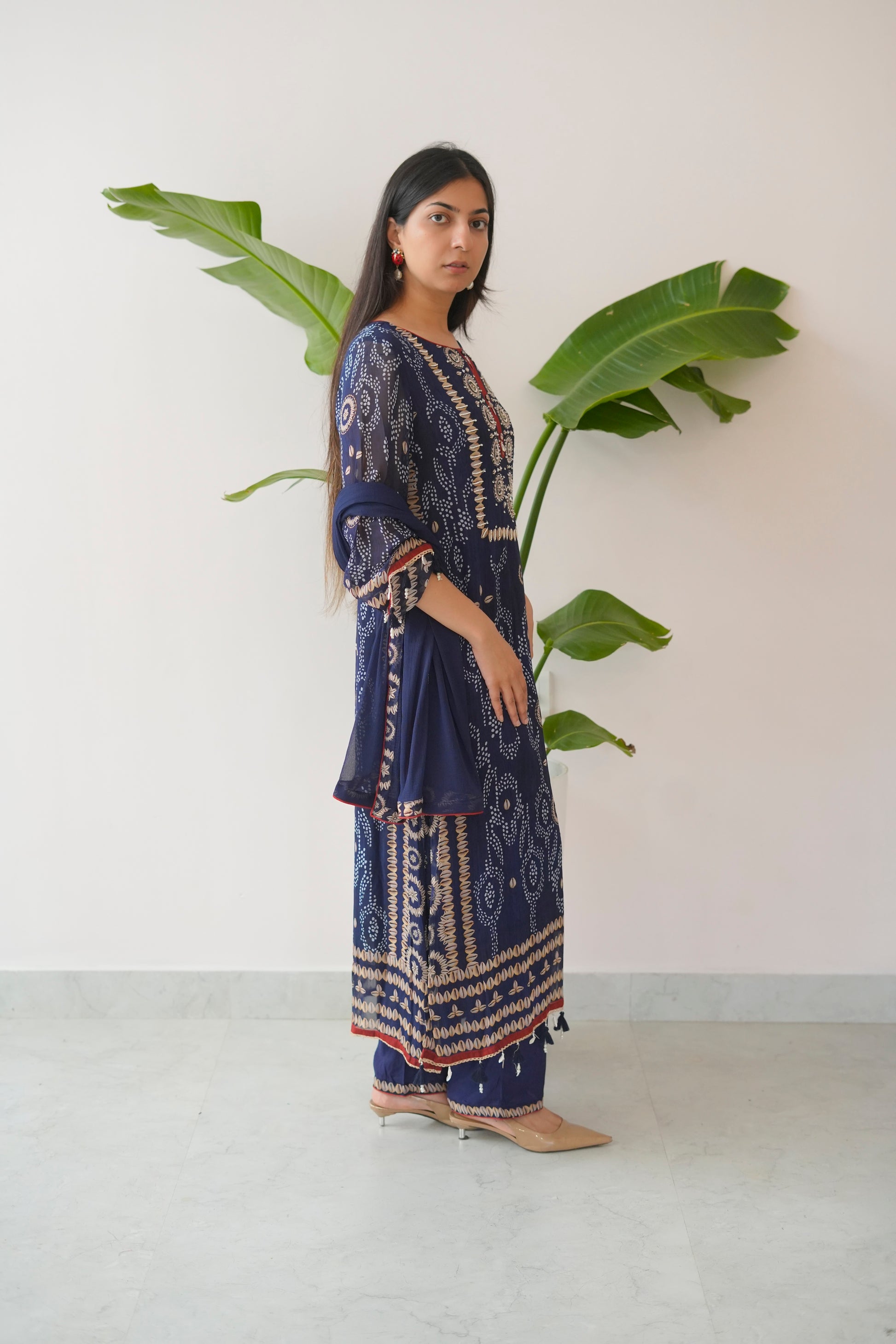 Woman in a blue patterned dress standing next to a large green leafy plant against a white wall.