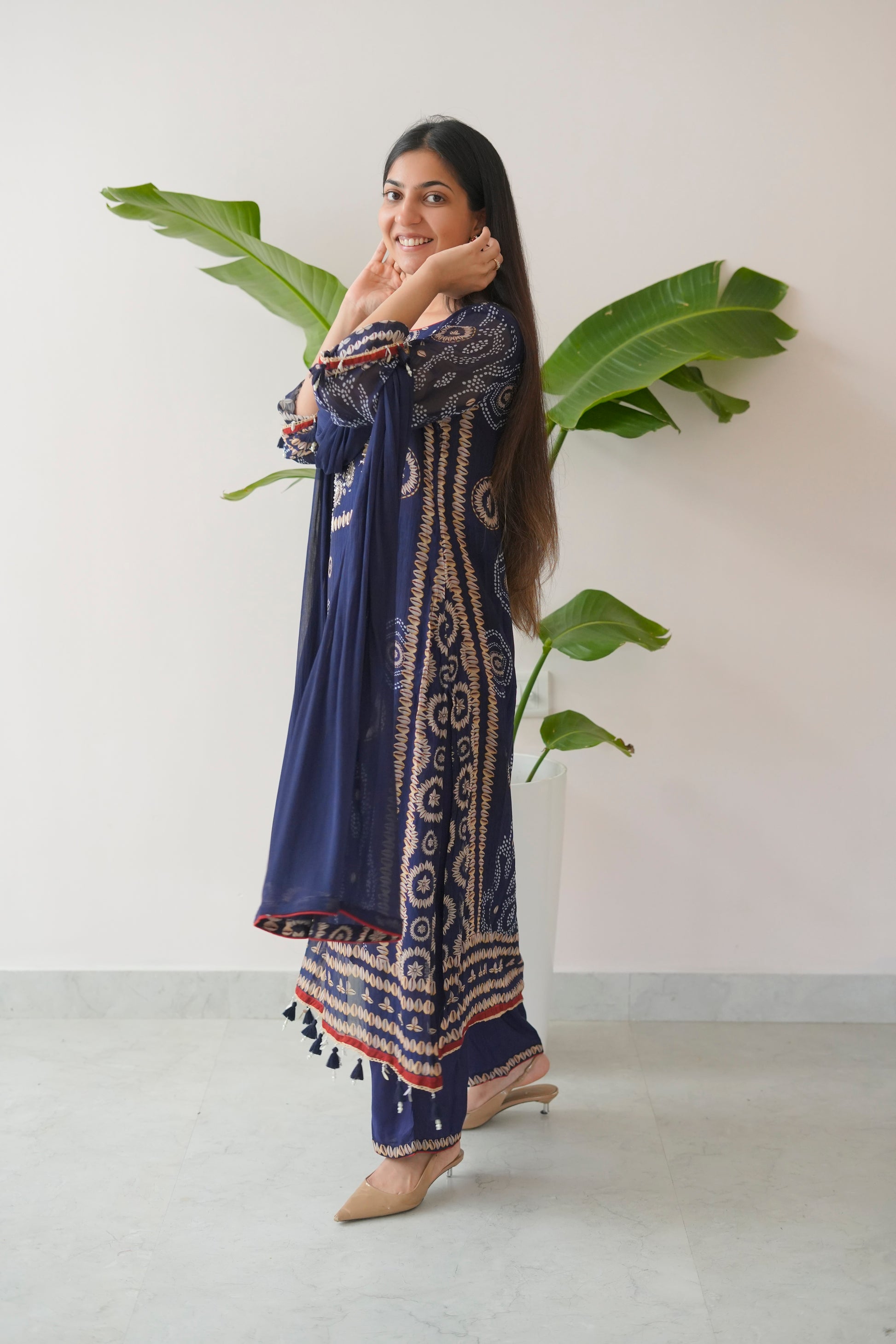 Woman wearing a blue patterned outfit with tassels, standing against a white wall with green leaves.