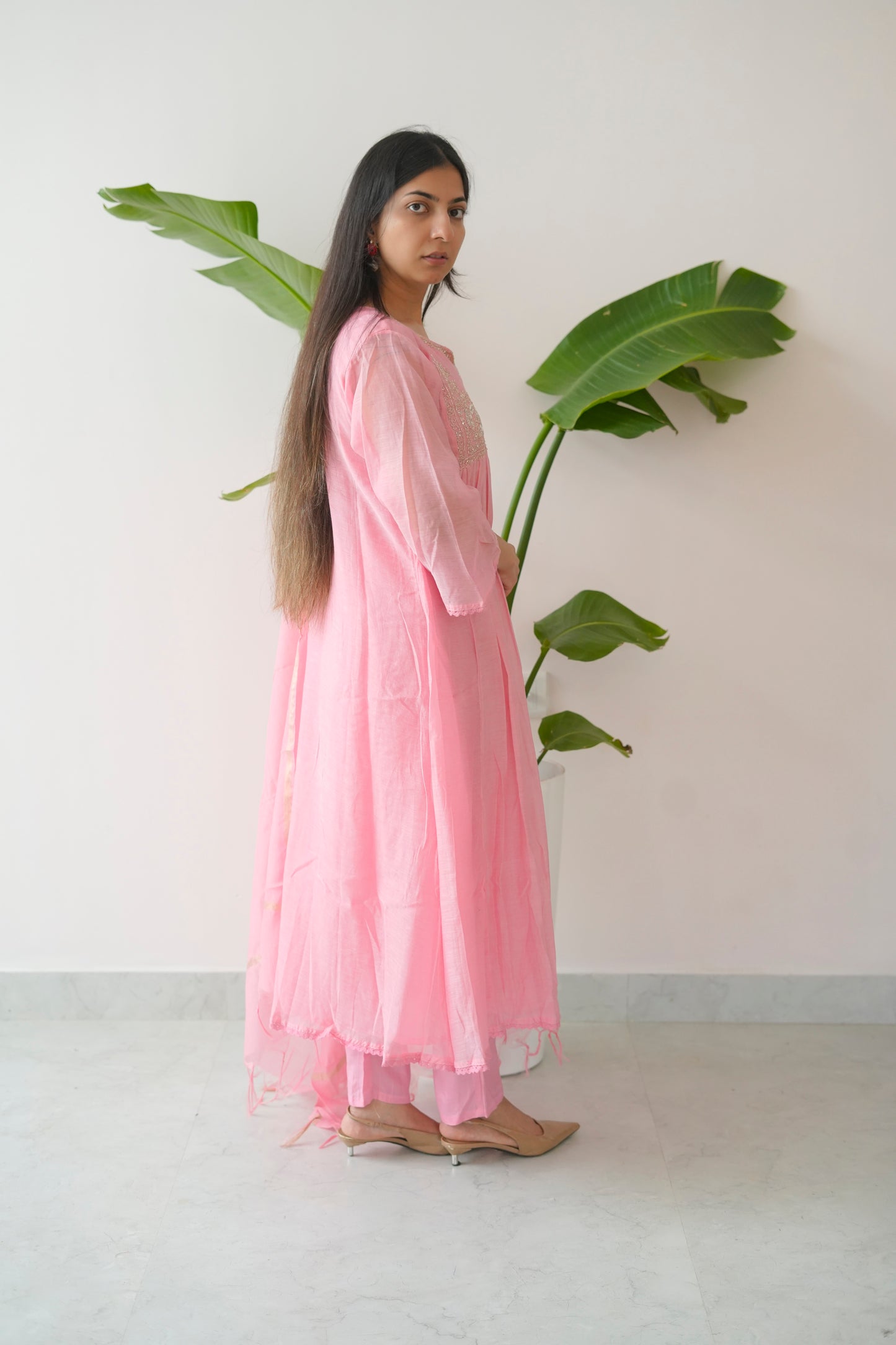 Woman in a pink dress holding a large green leaf against a white wall.