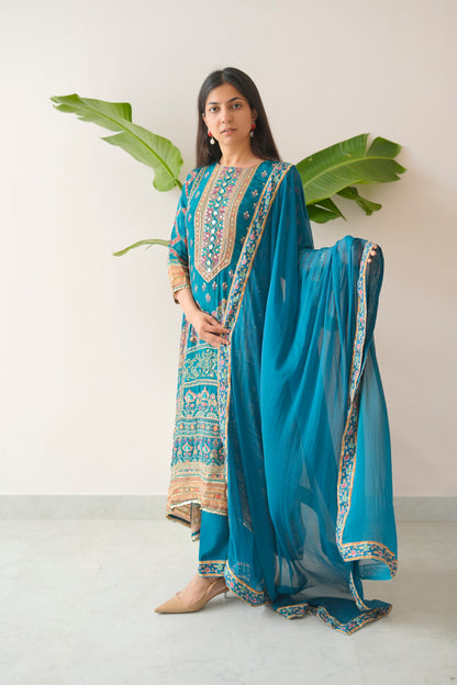 Woman wearing a blue embroidered outfit with a matching dupatta, standing against a white background with green leaves.