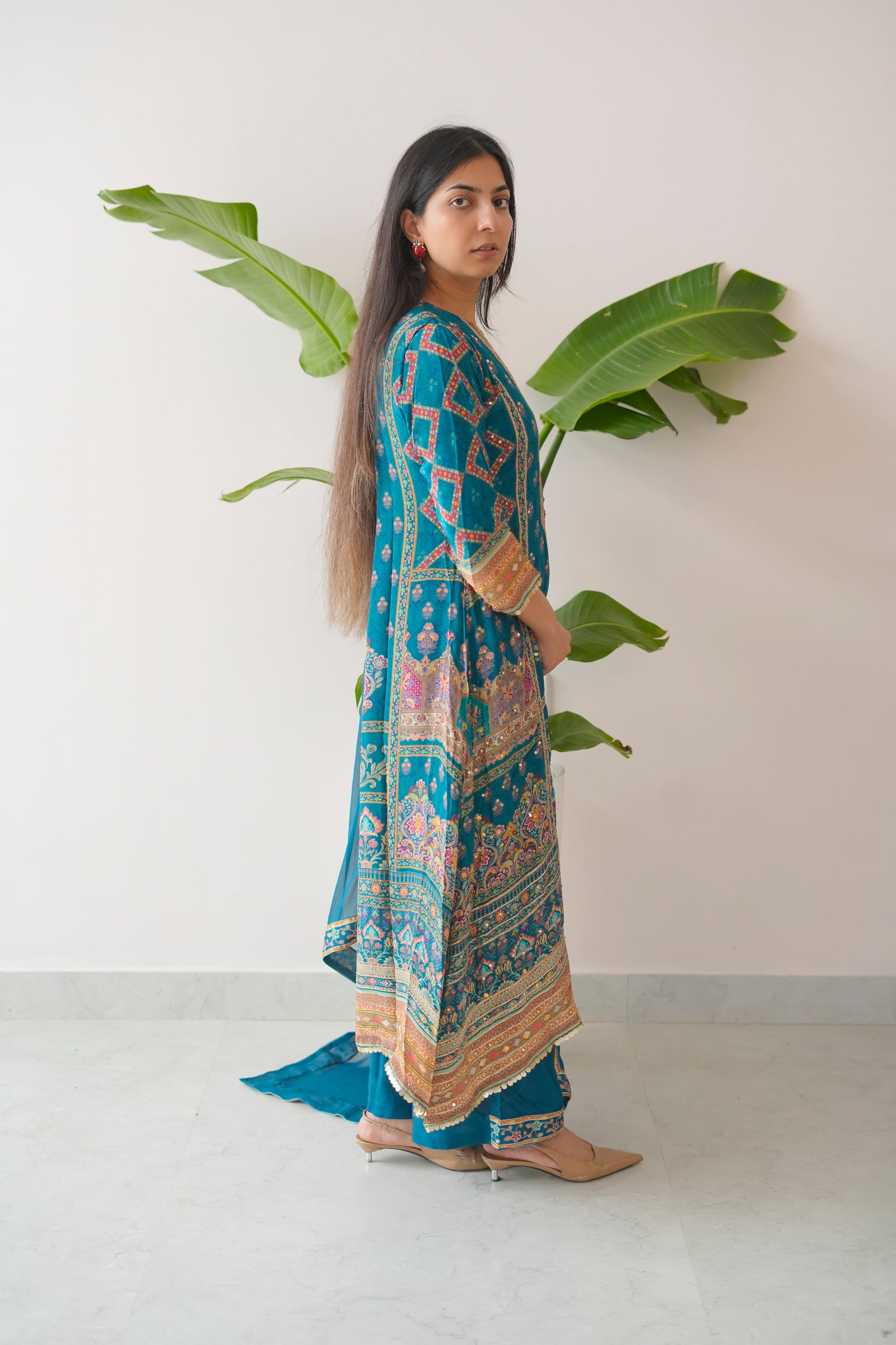 Woman wearing a blue patterned outfit standing against a white wall with green leaves.