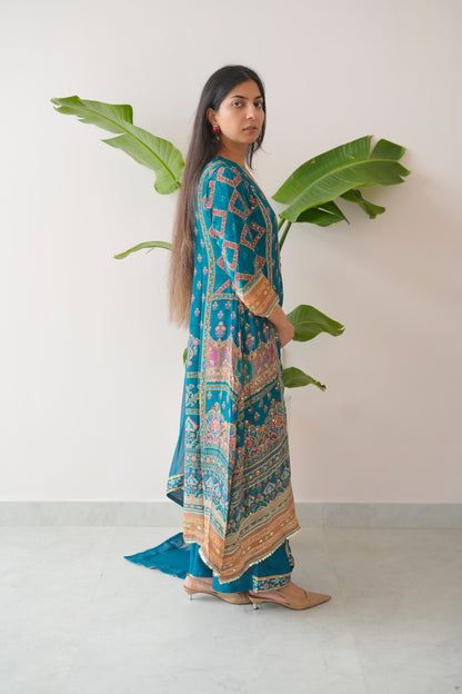 Woman wearing a blue patterned outfit standing against a white wall with green leaves.