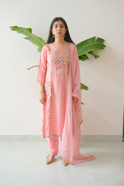 Woman wearing a pink traditional outfit with embroidery, standing against a white wall with green leaves.