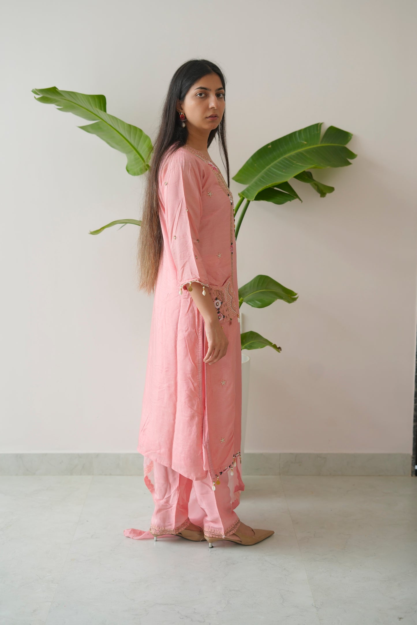 Woman in a pink outfit standing next to a large green leafy plant indoors.