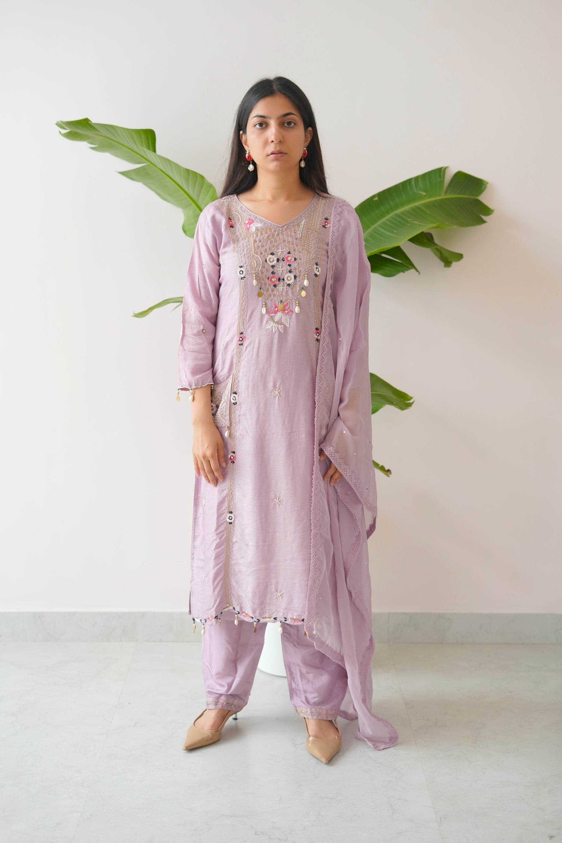 Woman wearing a light purple traditional outfit with floral embroidery, standing against a white background with green leaves.