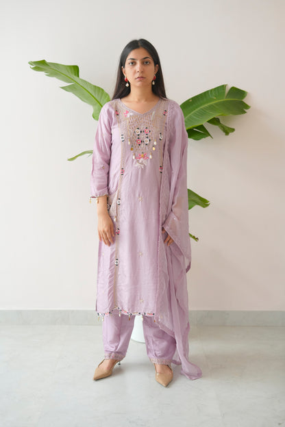 Woman wearing a light purple traditional outfit with floral embroidery, standing against a white background with green leaves.