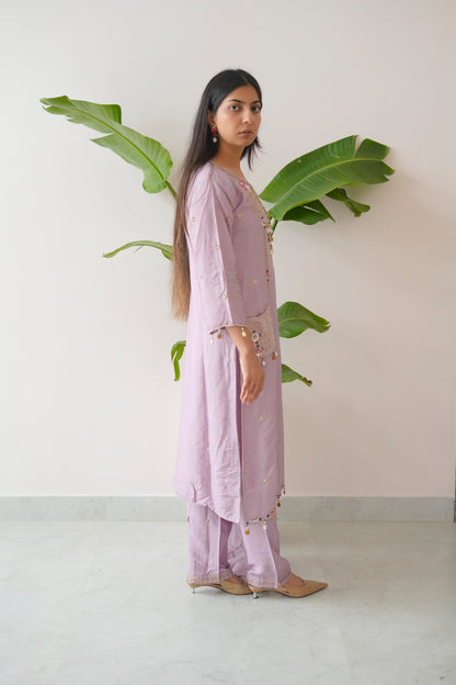 Woman in a light purple outfit standing in front of a white wall with green leaves.