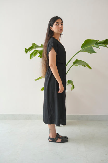 Woman wearing a black dress standing next to a plant indoors.