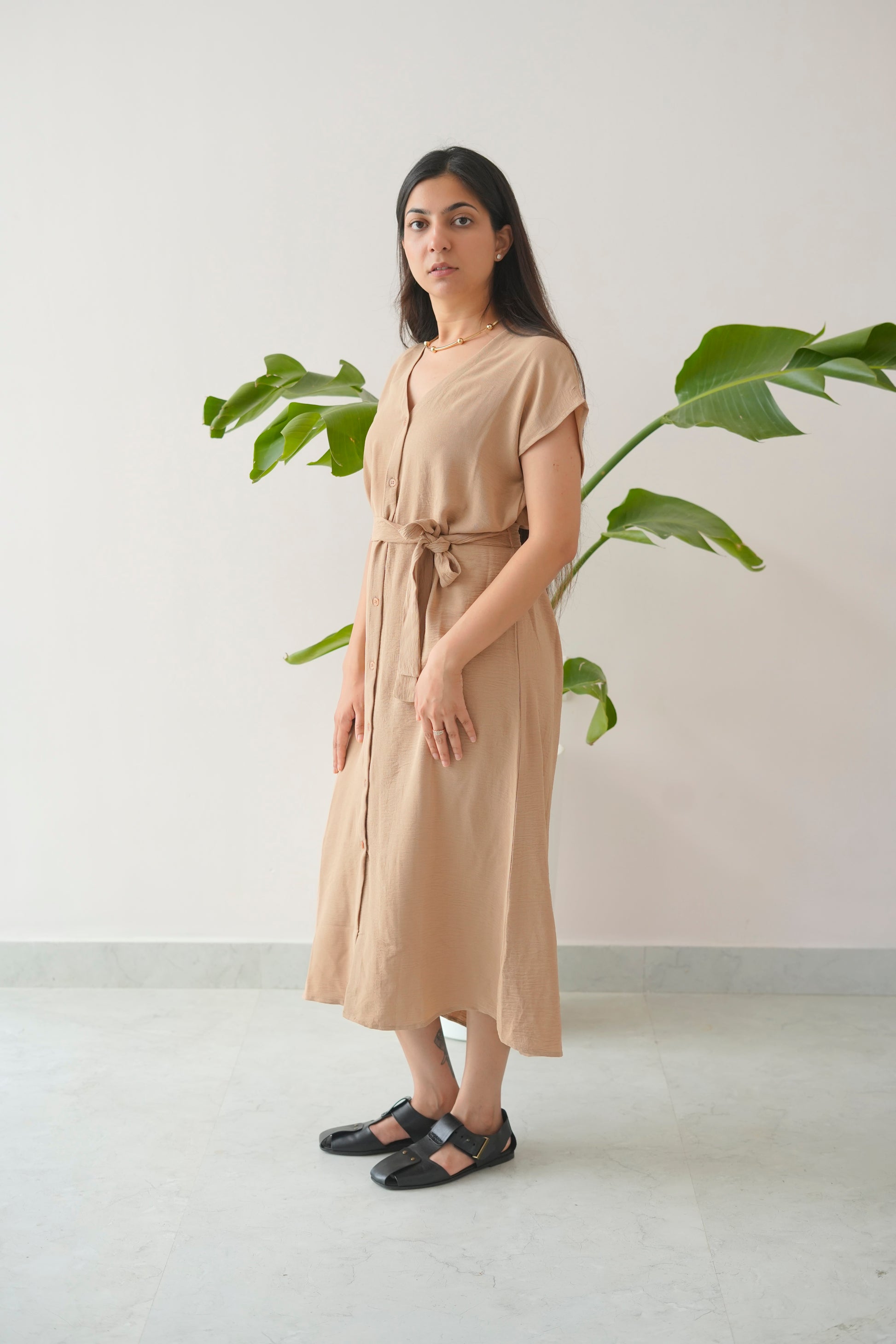 Woman wearing a beige dress standing in front of a plant against a white wall.