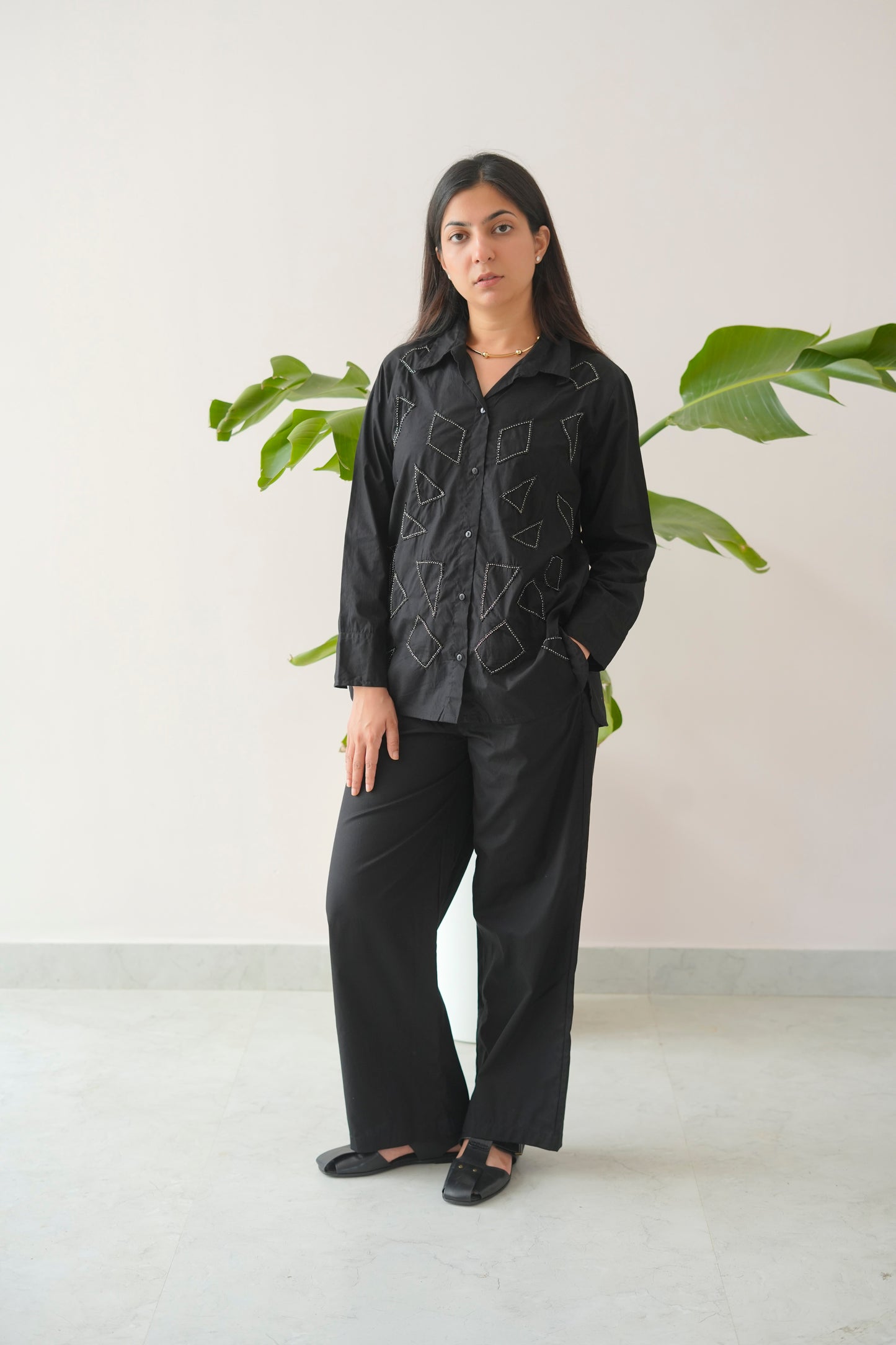 Woman wearing a black outfit with embroidery in a minimalistic setting with plants.