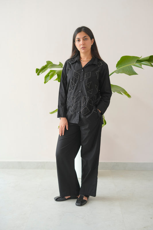 Woman wearing a black outfit with embroidery in a minimalistic setting with plants.