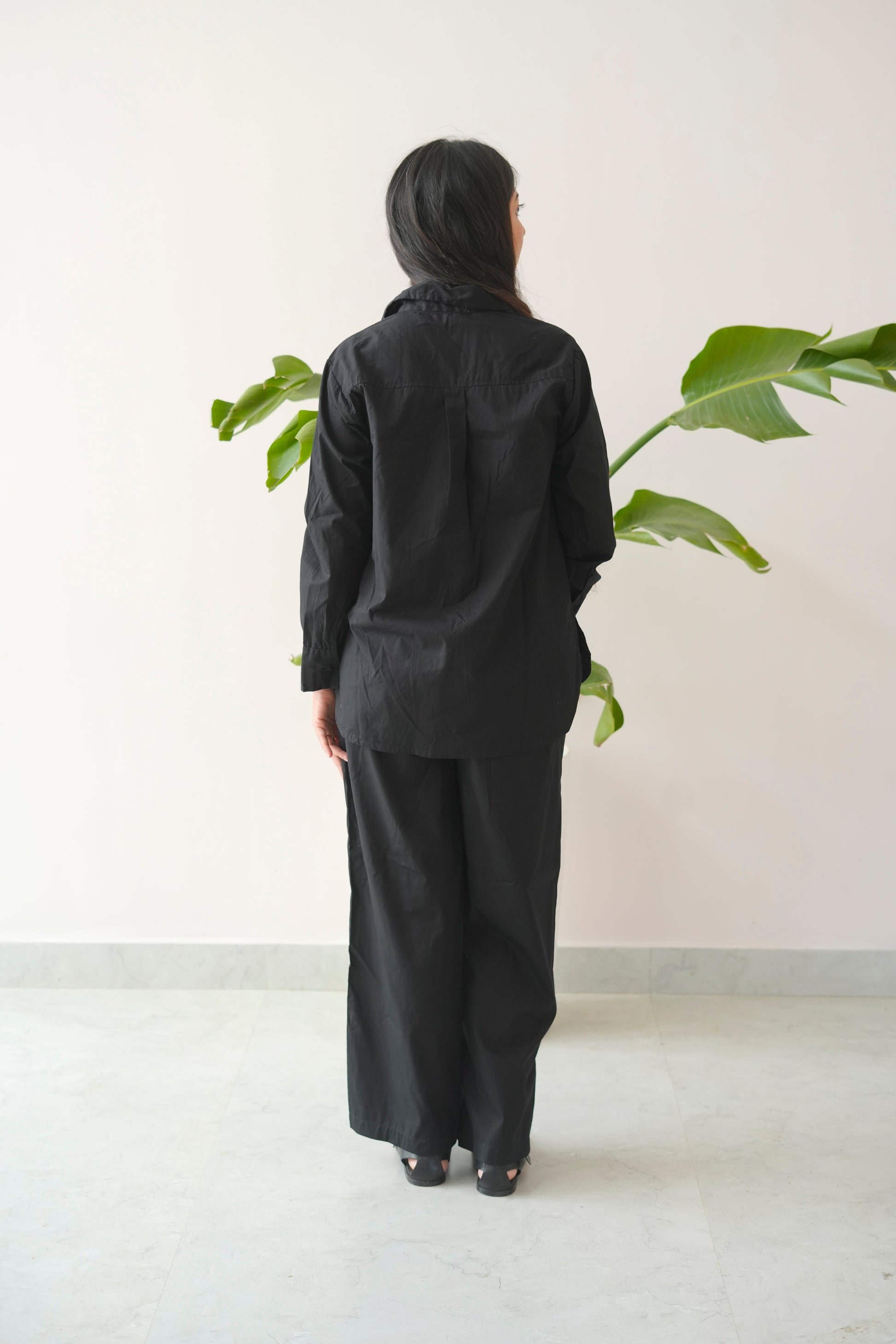 Person wearing a black outfit standing in front of a plant against a white wall.