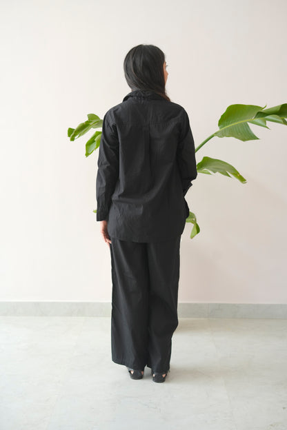 Person wearing a black outfit standing in front of a plant against a white wall.