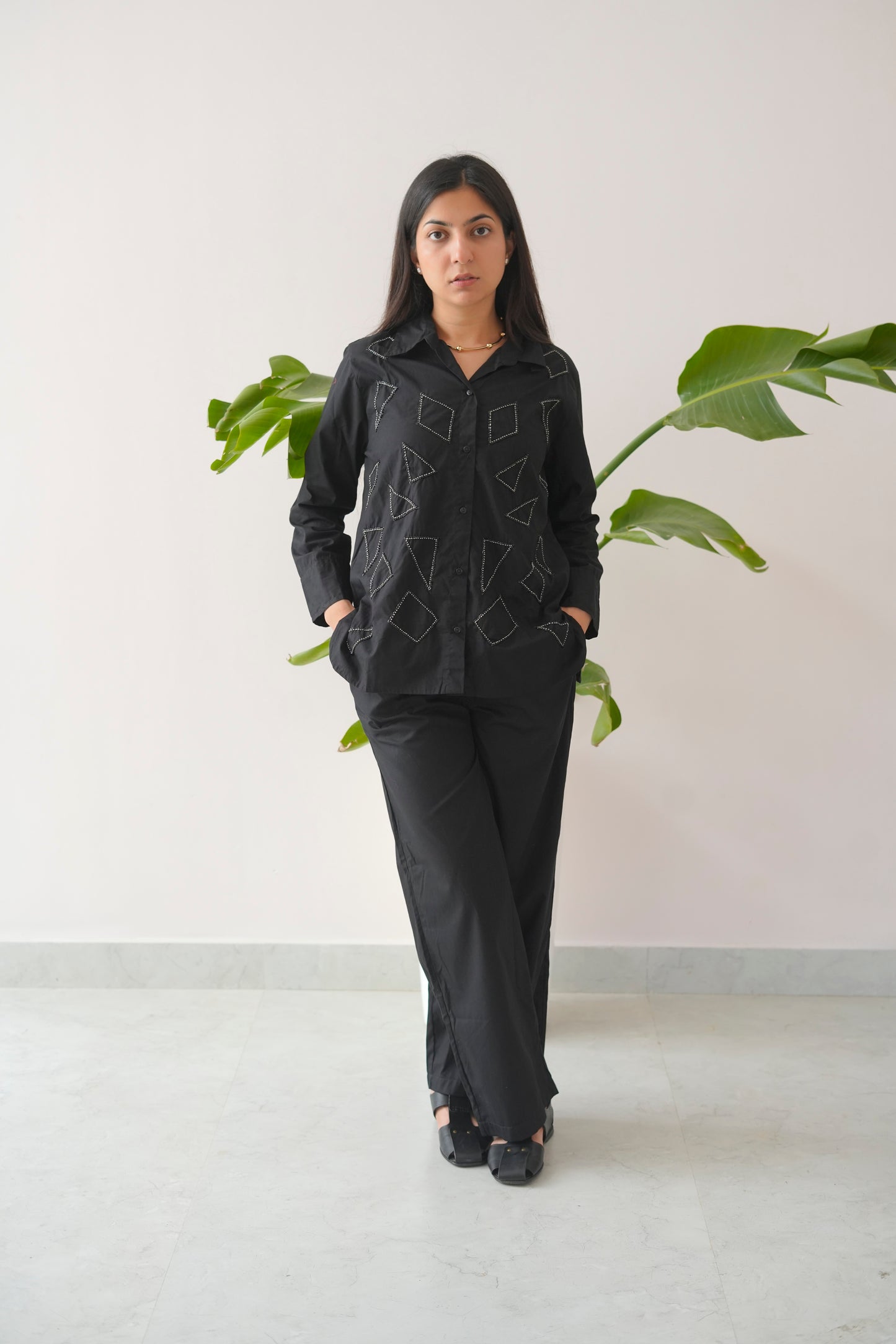 Woman wearing a dark gray jacket and pants standing in front of a white wall with a plant.