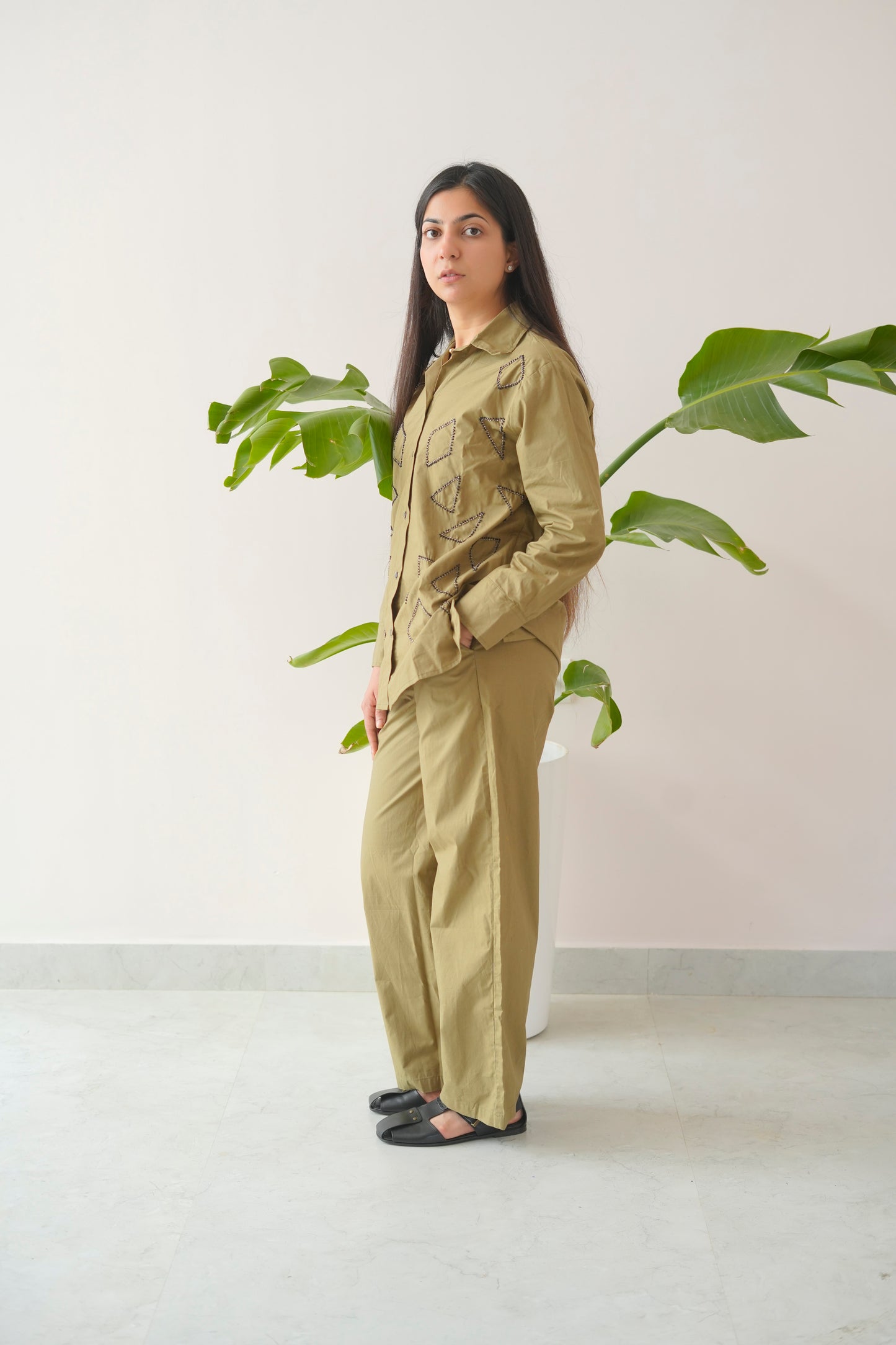 Person wearing a green jumpsuit standing next to a plant against a white wall.