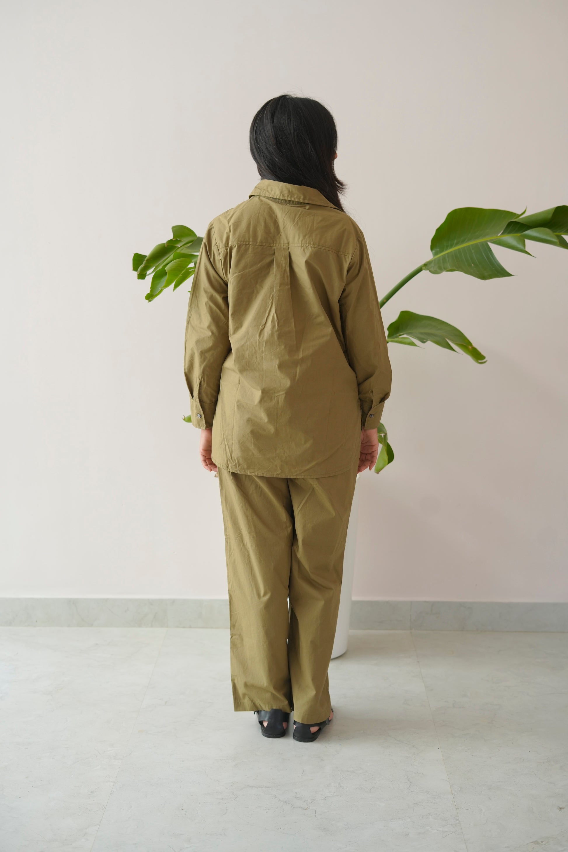 Person wearing a green outfit standing in front of a plant against a white wall.