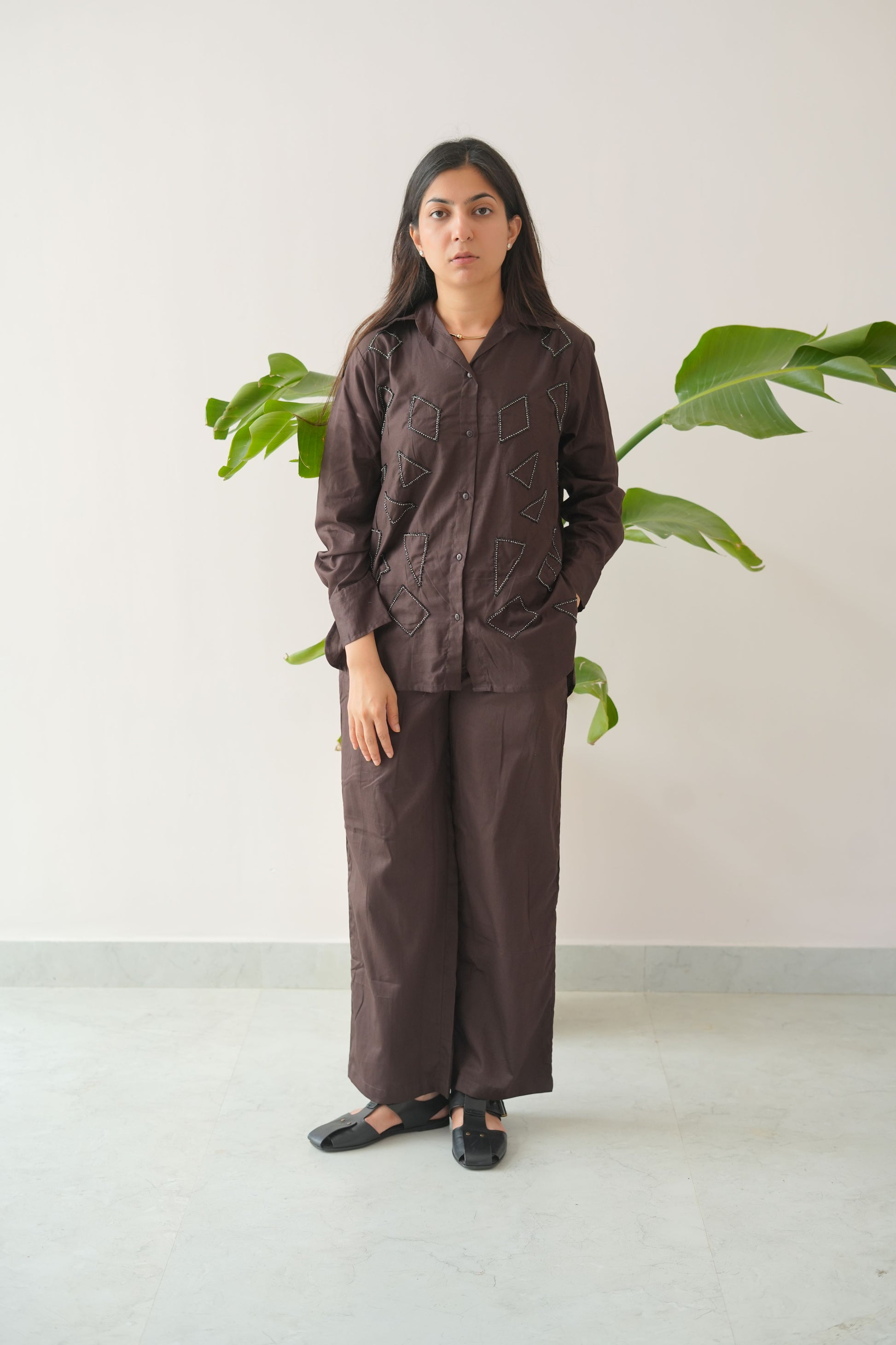 Woman wearing a brown outfit standing against a white wall with a plant in the background