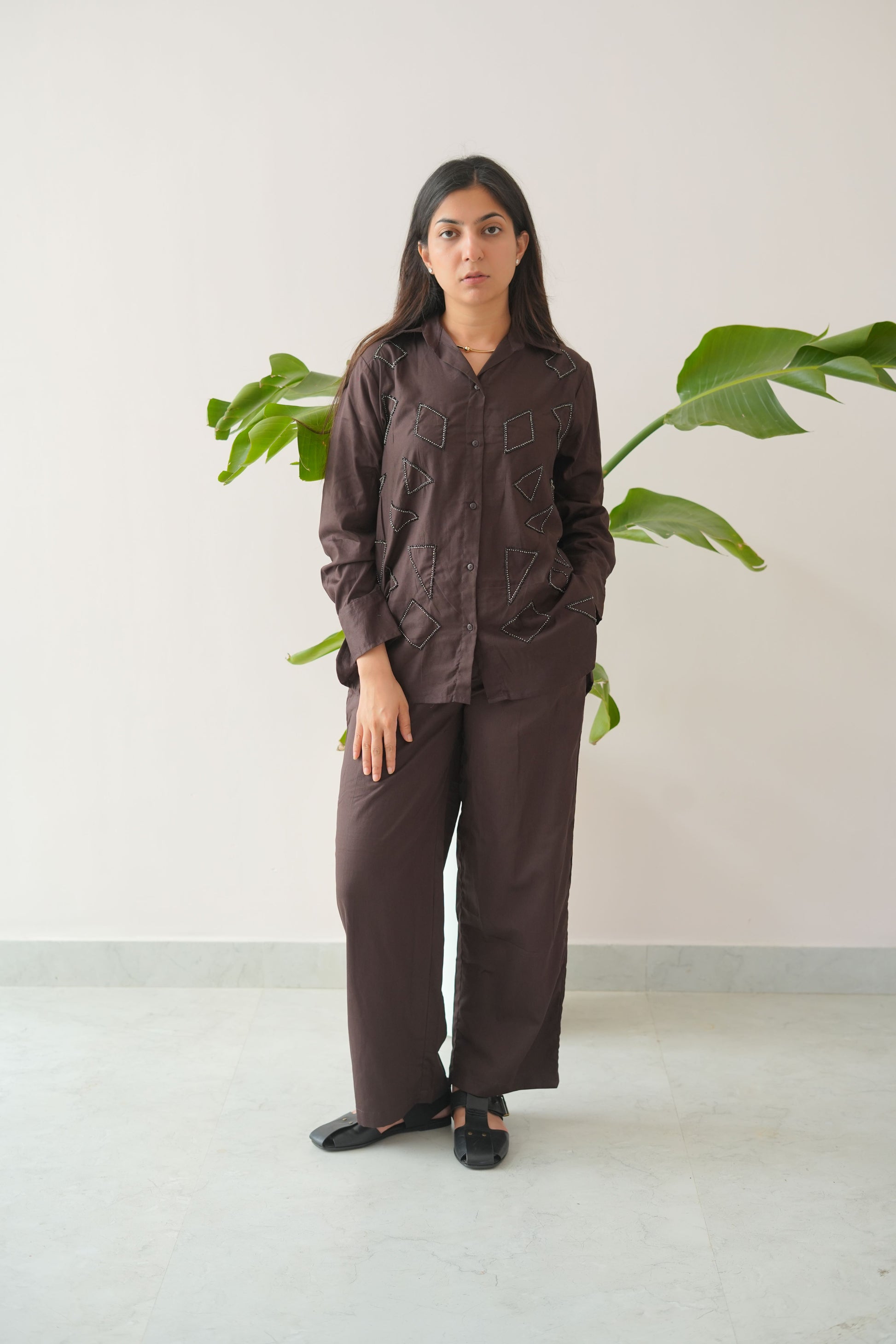 Person wearing a brown outfit standing against a light background with green plants.