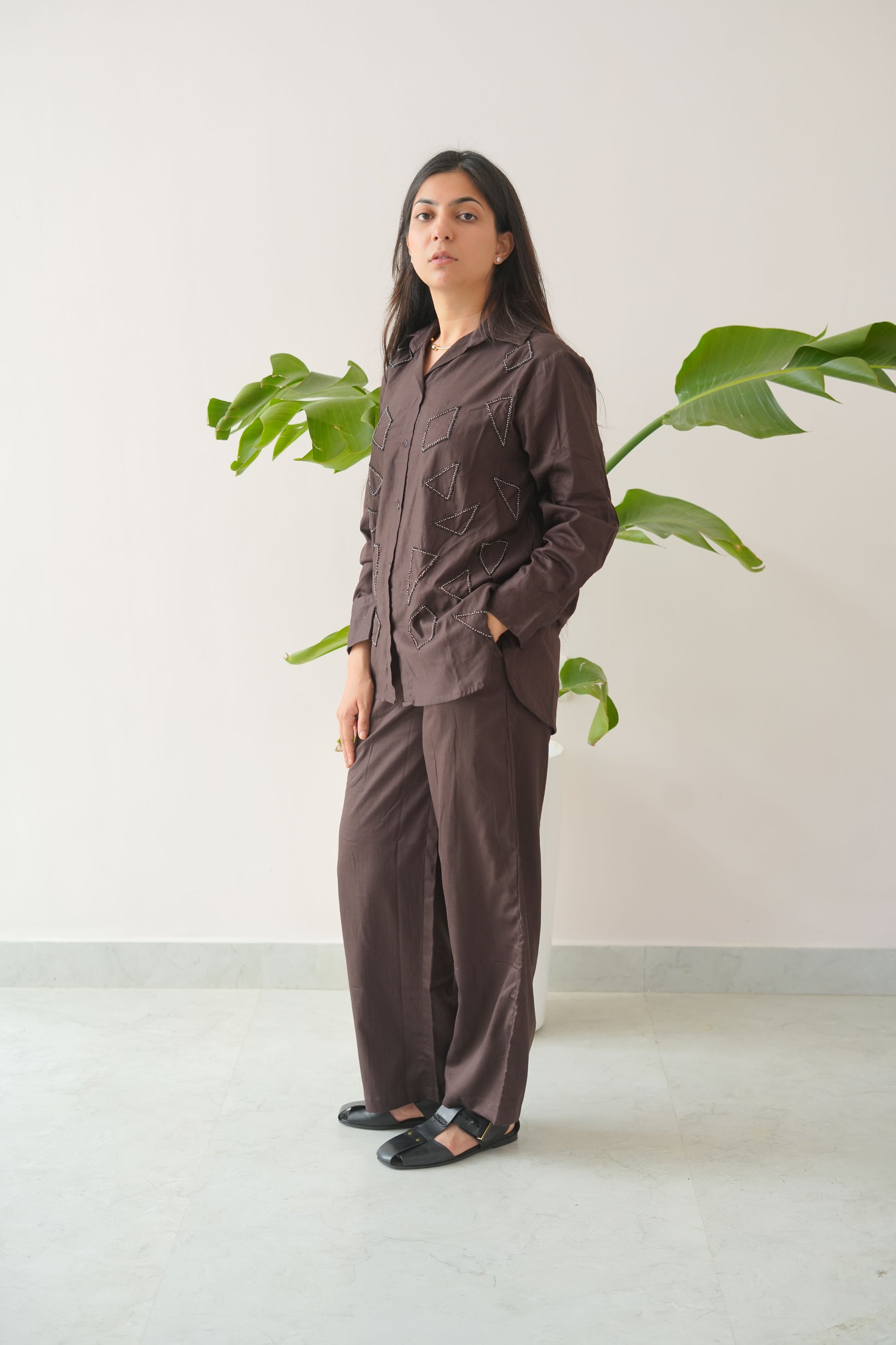 Woman in a brown dress standing against a white wall with green plants.