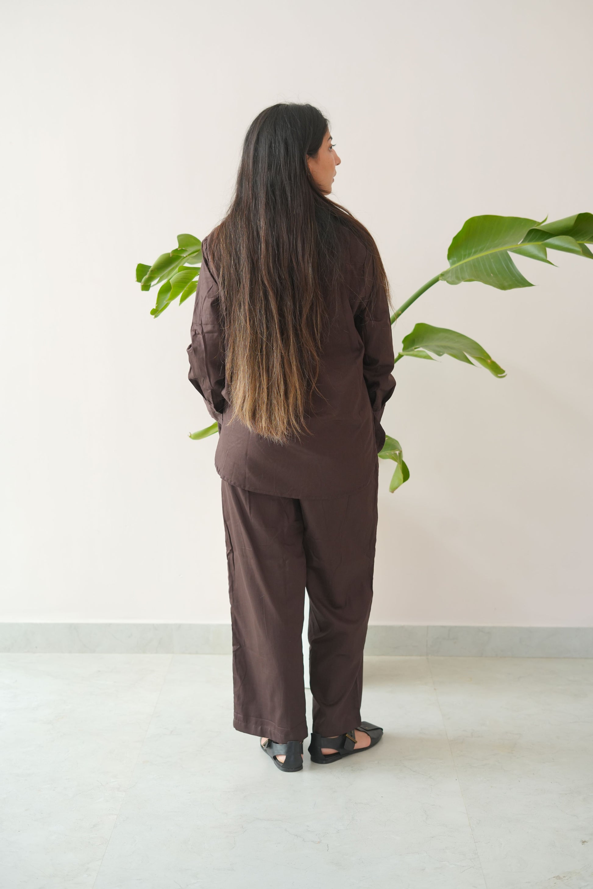 Person wearing a brown outfit standing against a white wall with a plant.