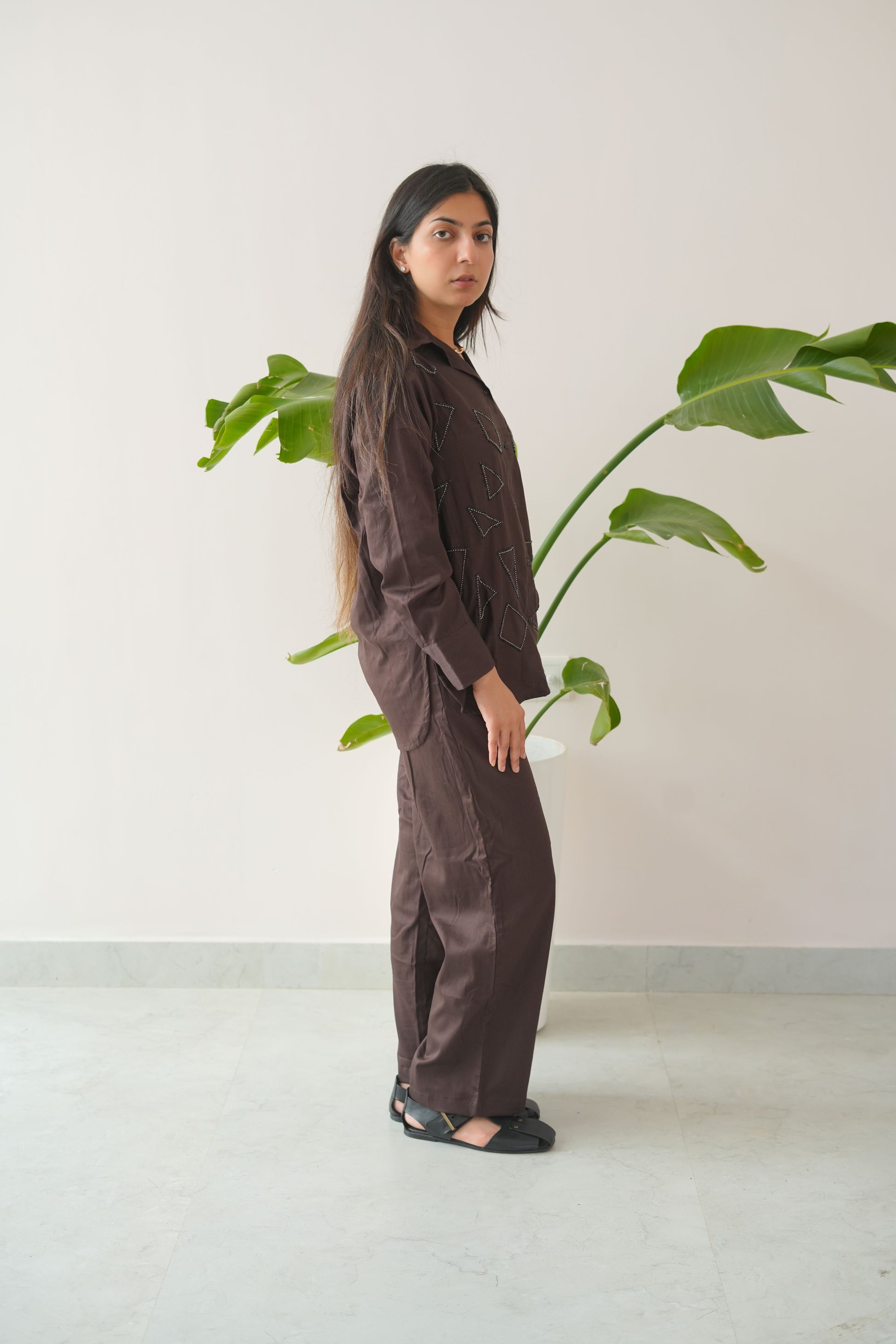 Person wearing a dark outfit standing next to a plant against a white wall.