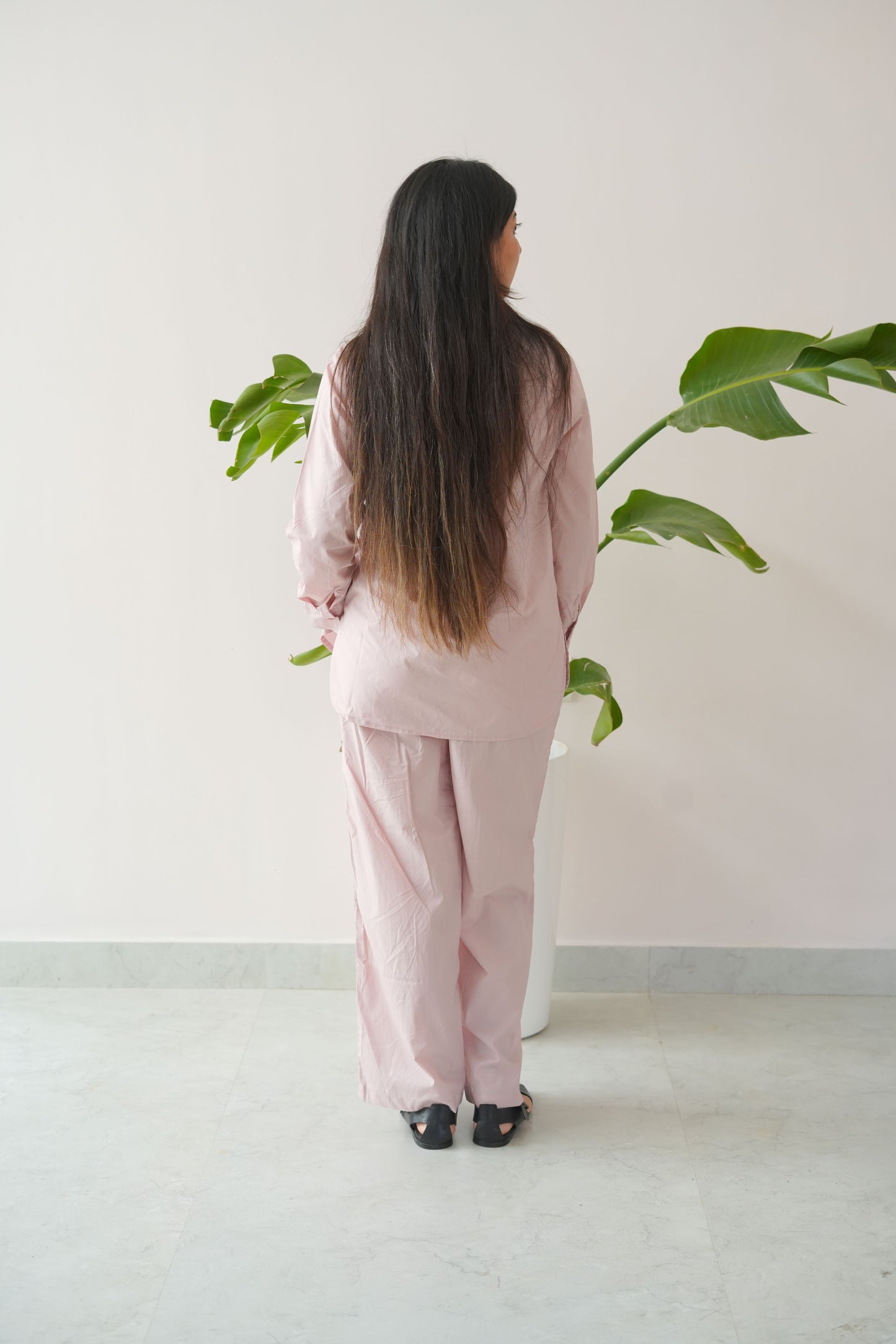 Person wearing a pink outfit standing against a white wall with green plants.