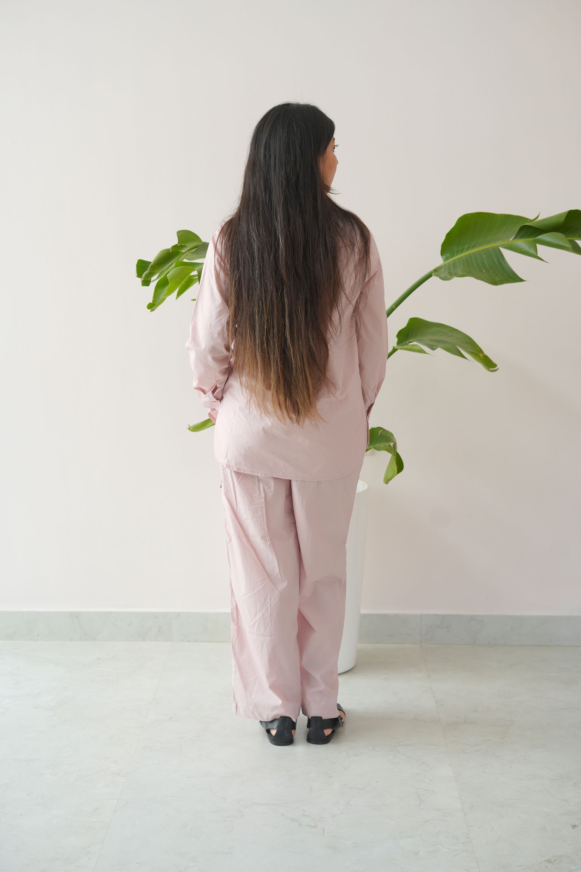 Person wearing a pink outfit standing against a white wall with green plants.