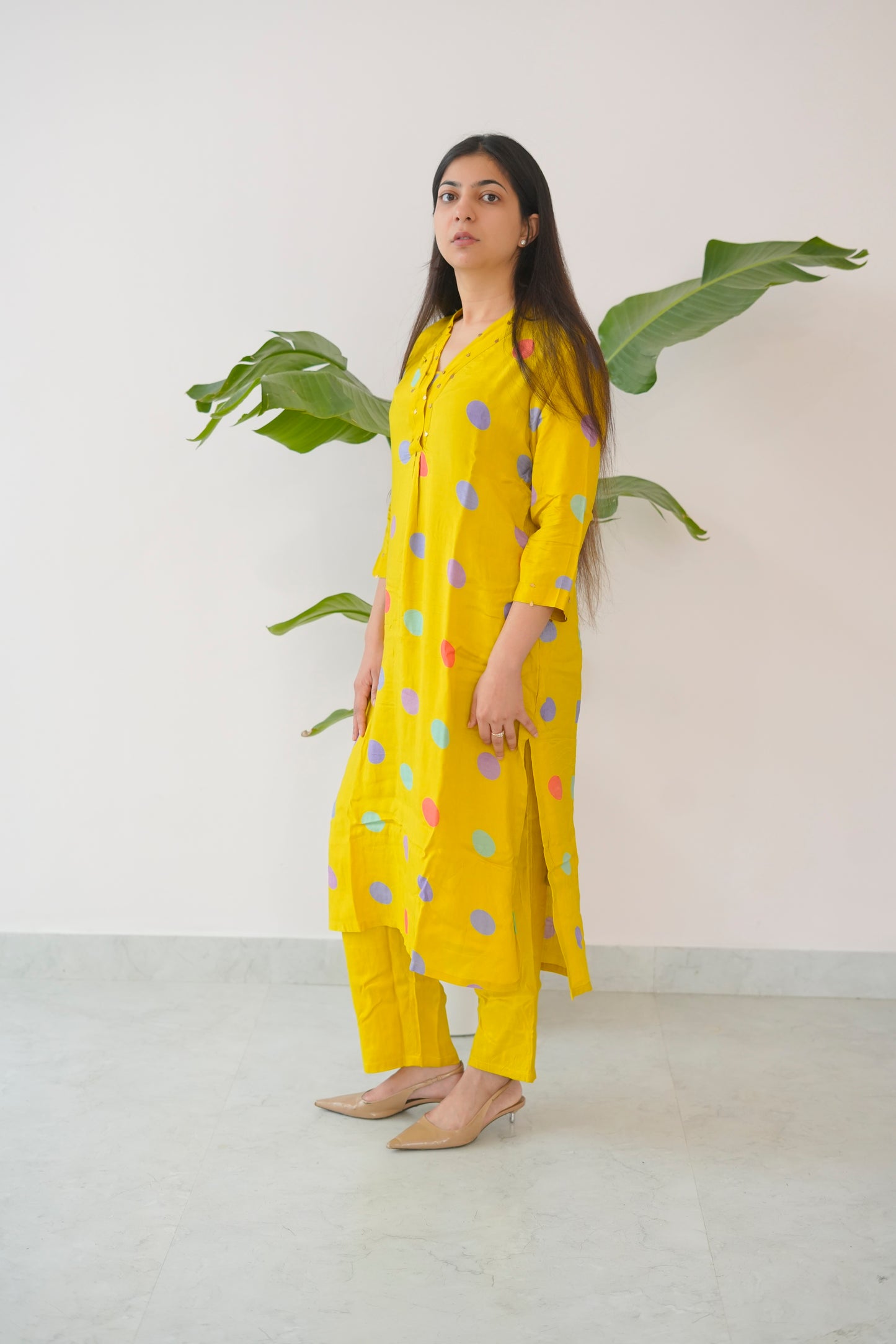 Woman wearing a yellow polka dot dress standing against a white wall with green leaves.