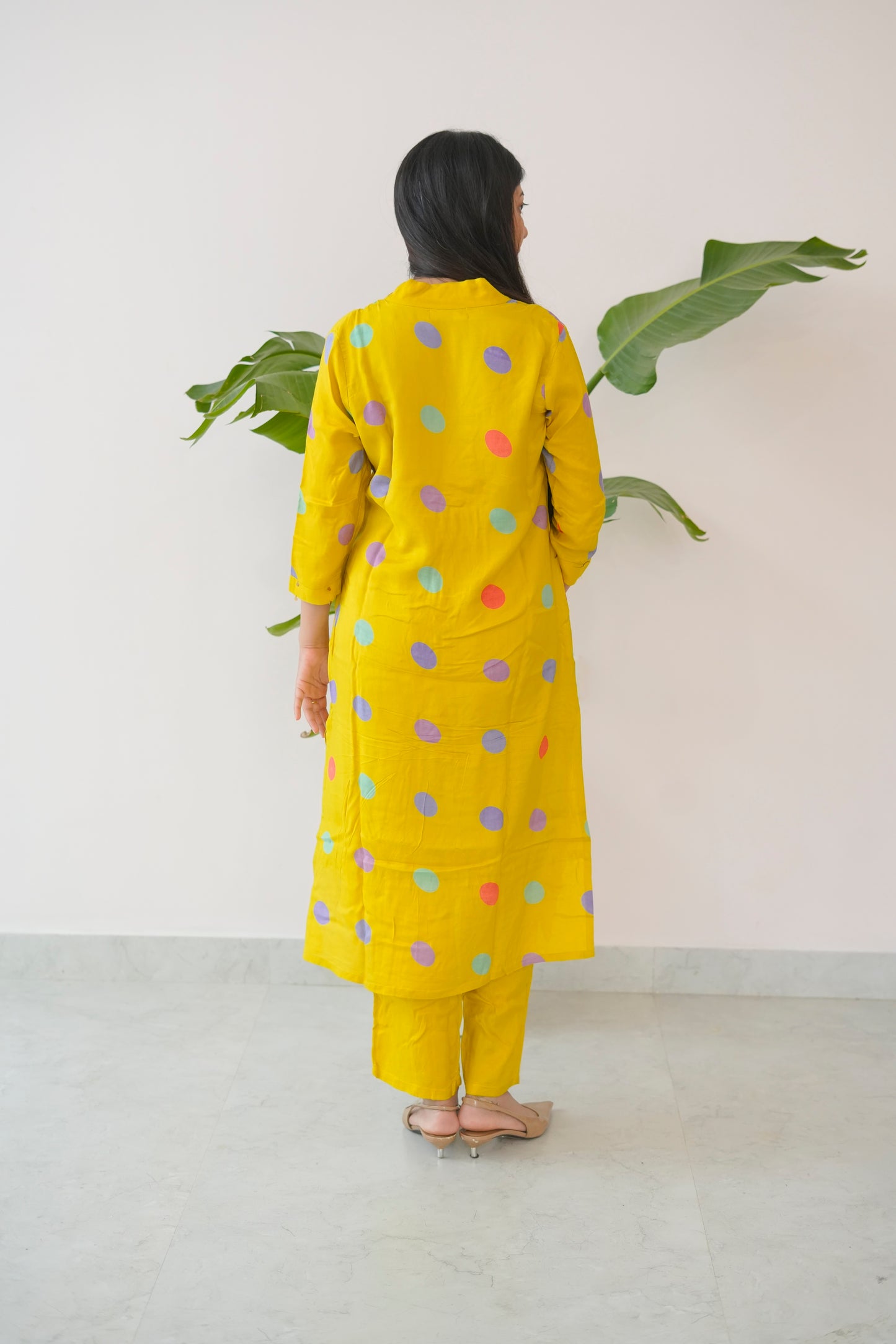 Person wearing a yellow outfit with colorful polka dots, standing against a white wall with green leaves.