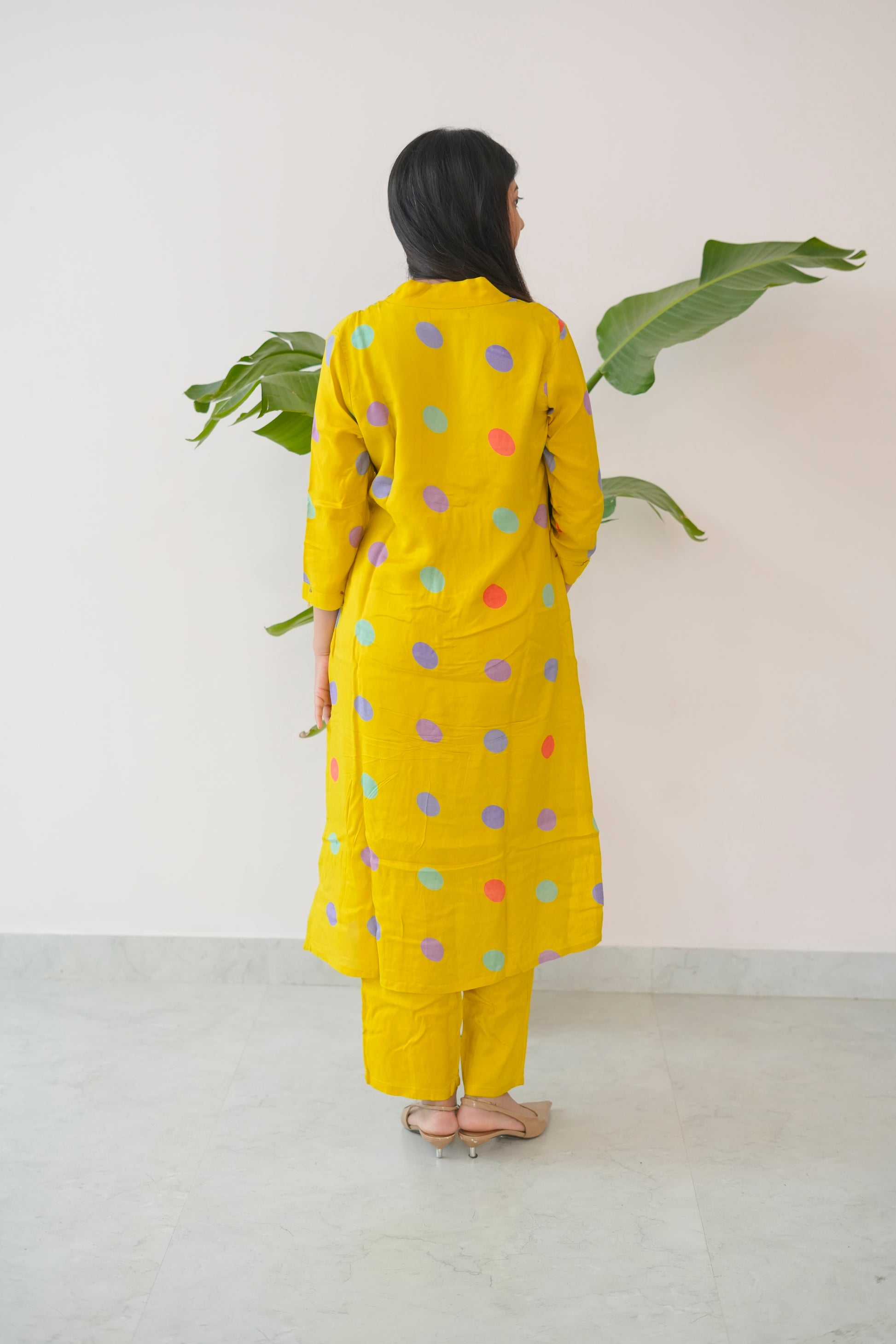 Person wearing a yellow outfit with colorful polka dots, standing against a white wall with green leaves.