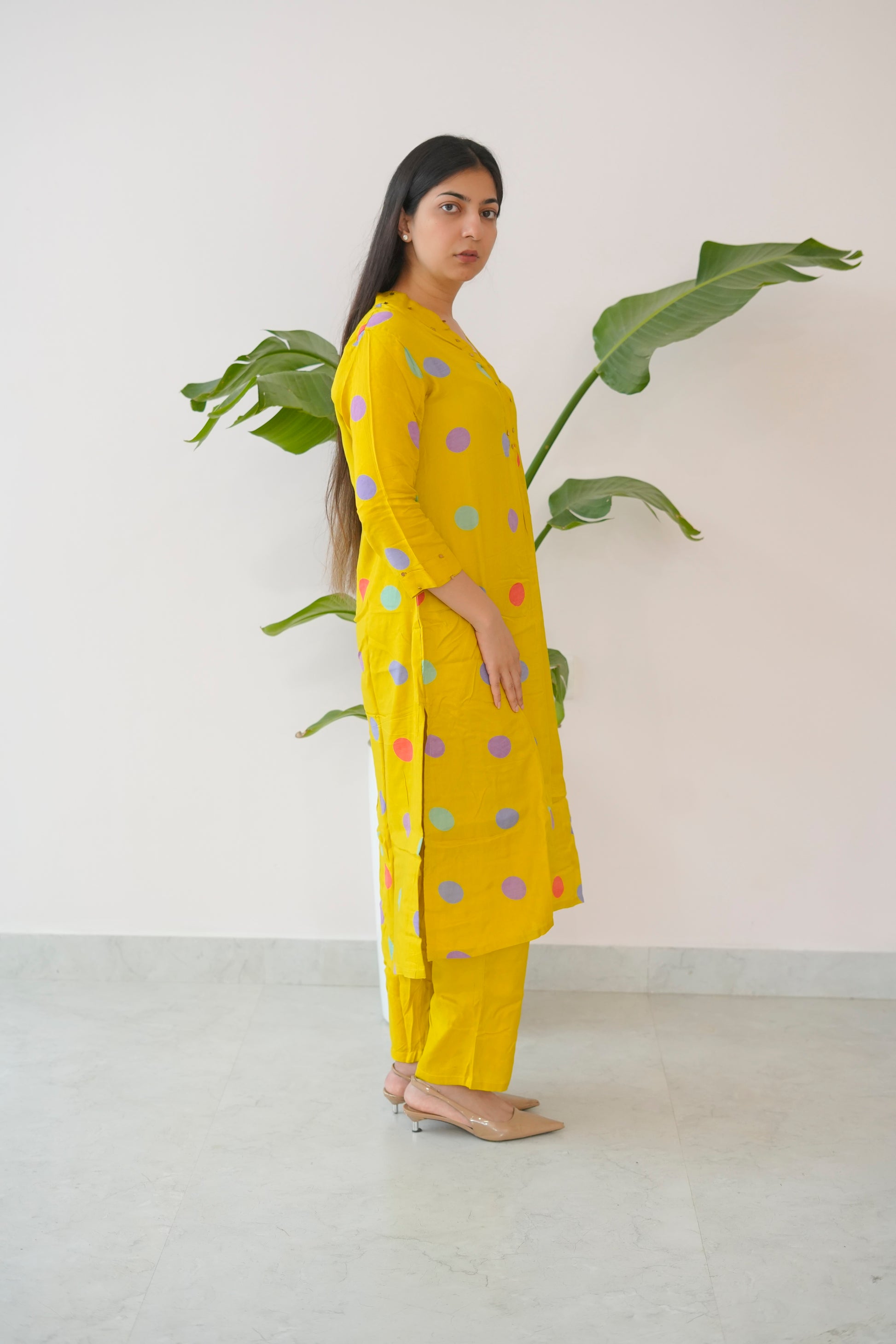 Woman wearing a yellow outfit with colorful polka dots standing next to a plant against a white wall.