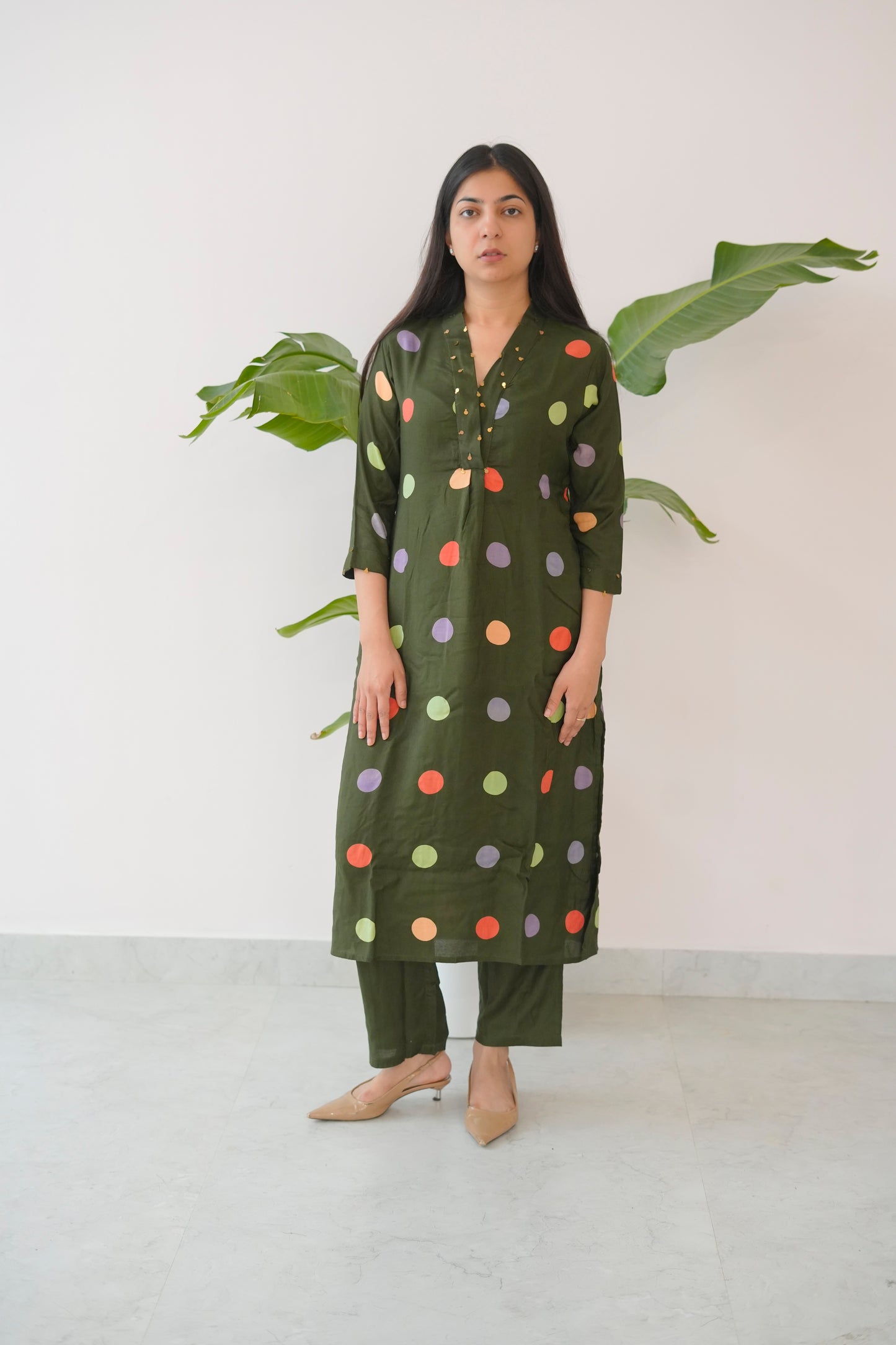 Woman wearing a green polka dot dress standing in front of a white wall with a plant.
