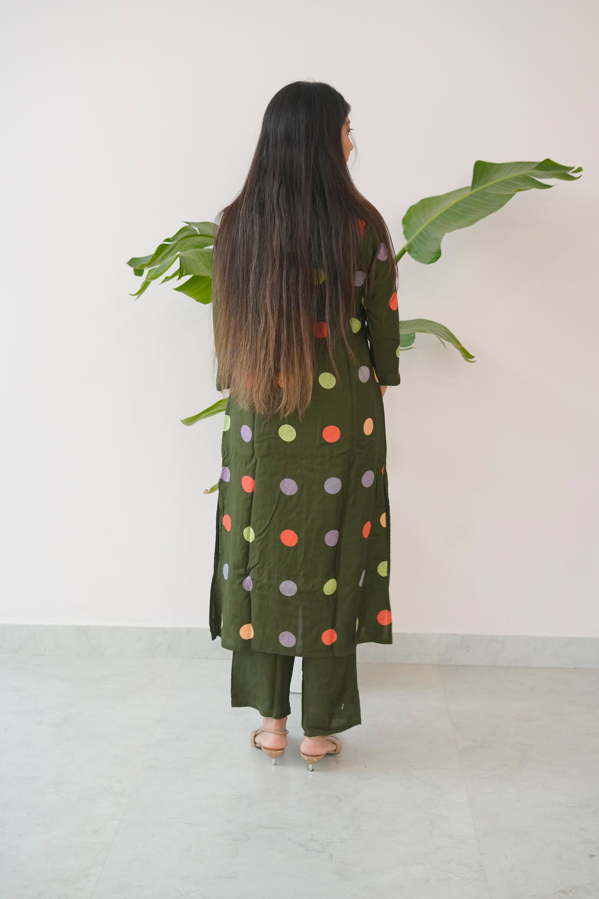 Woman wearing a green polka dot outfit standing in front of a white wall with a plant.