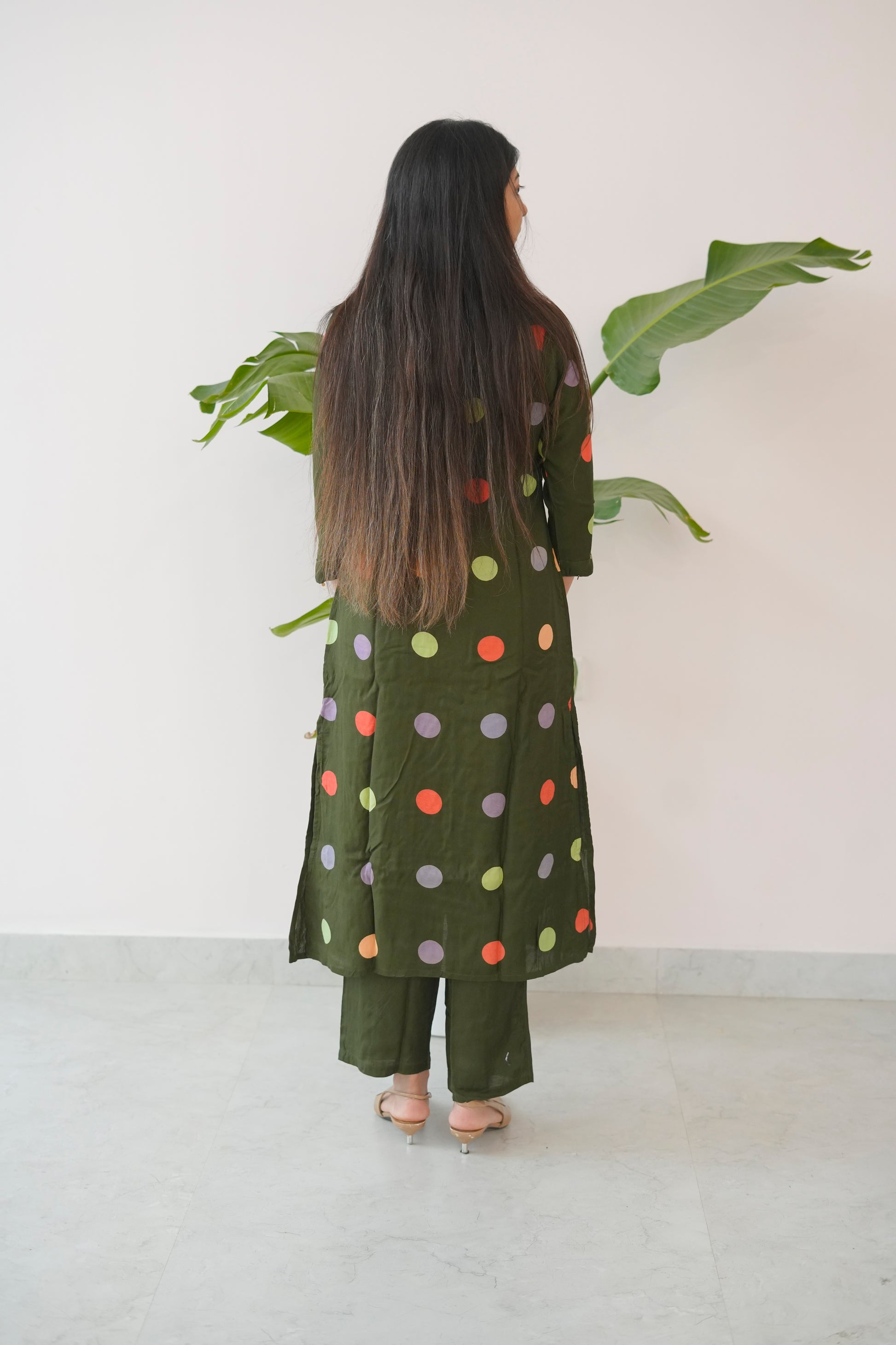 Person wearing a green outfit with colorful polka dots, standing in front of a white wall with a plant.
