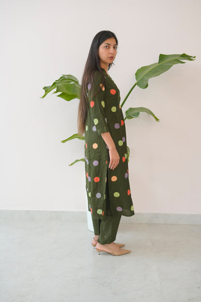 Woman wearing a green polka dot dress standing next to a plant indoors.