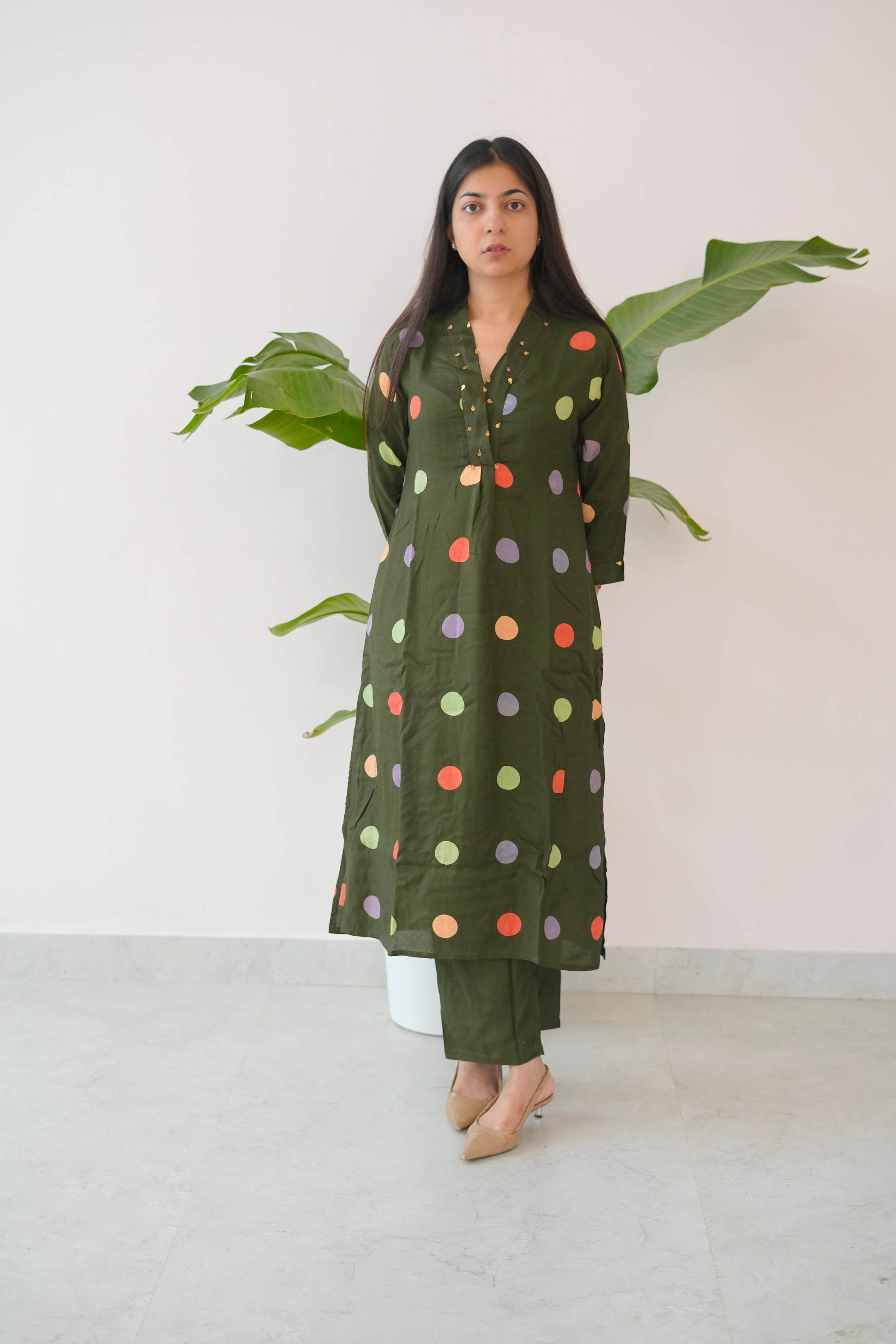 Woman wearing a green polka dot dress standing against a white wall with a plant.
