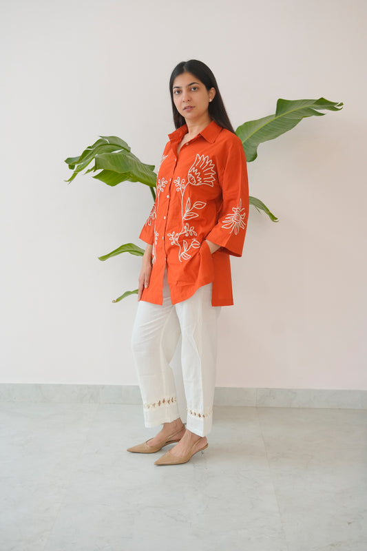 Woman wearing an orange embroidered shirt and white pants standing in front of a plant.