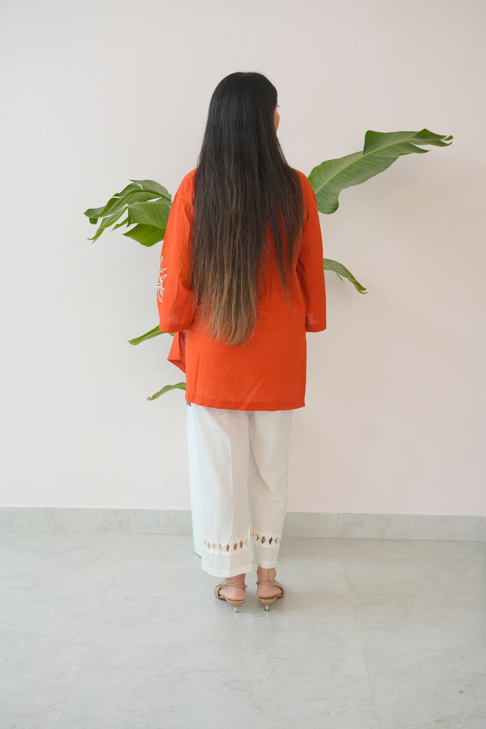 Person wearing an orange top and white pants standing in front of a plant against a plain wall.